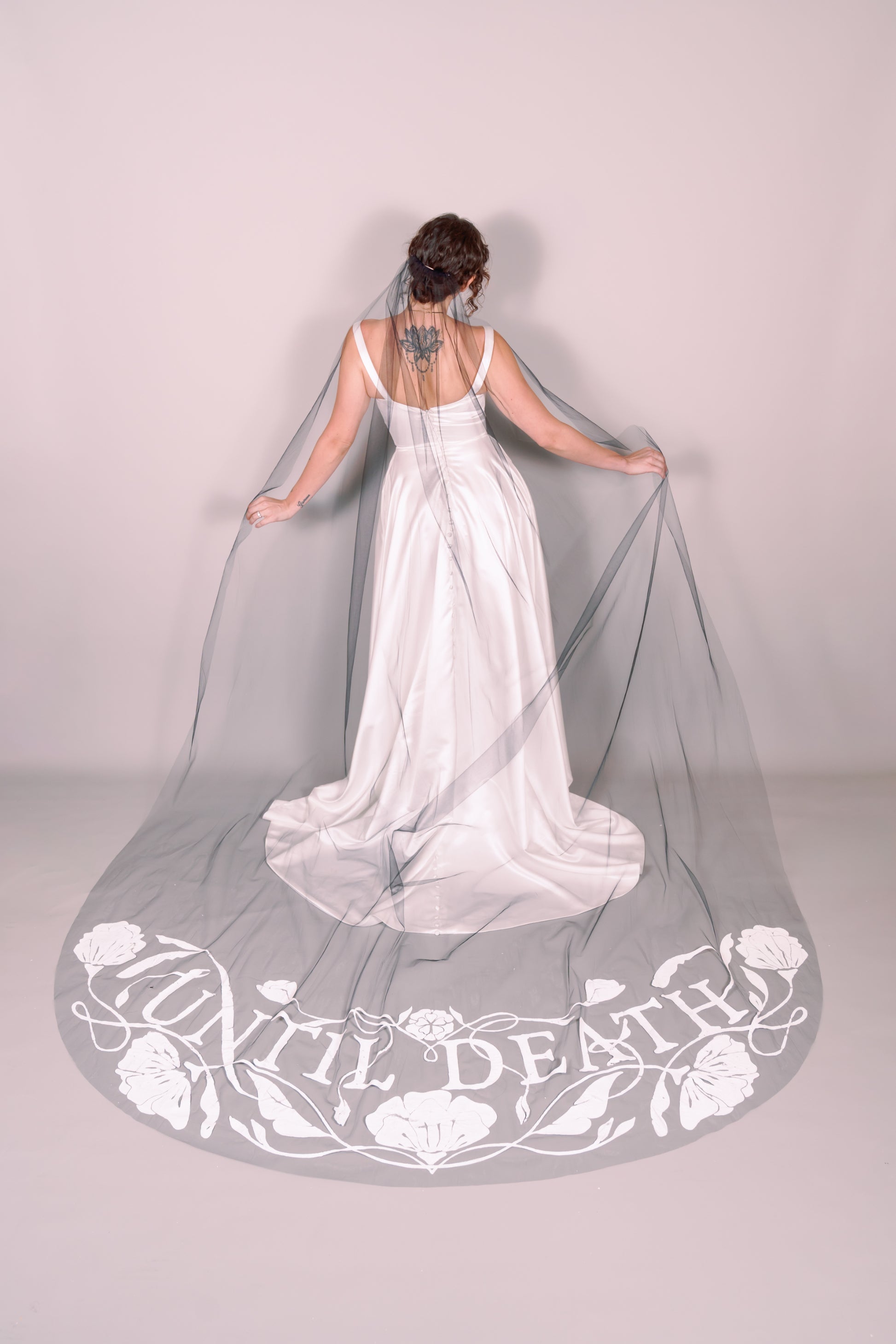 Woman in a white dress holding a long black veil with 'UNTIL DEATH' text on a plain background. Photography by Rachel Sakakeeny Model is Isabelle Paulino