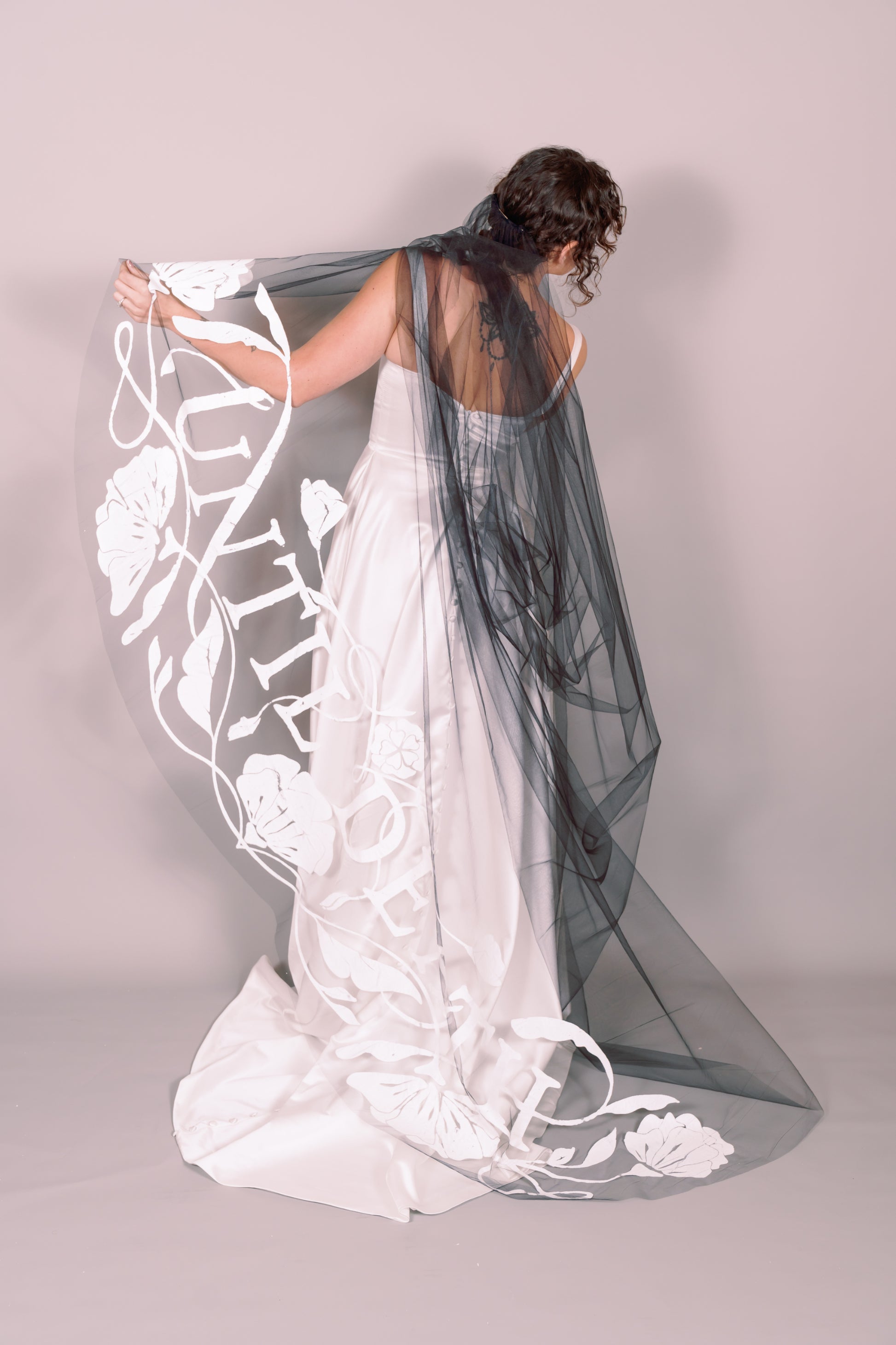 Woman in a white wedding dress with a long black painted veil that says Until Death against a plain background. Photography by Rachel Sakakeeny Model is Isabelle Paulino