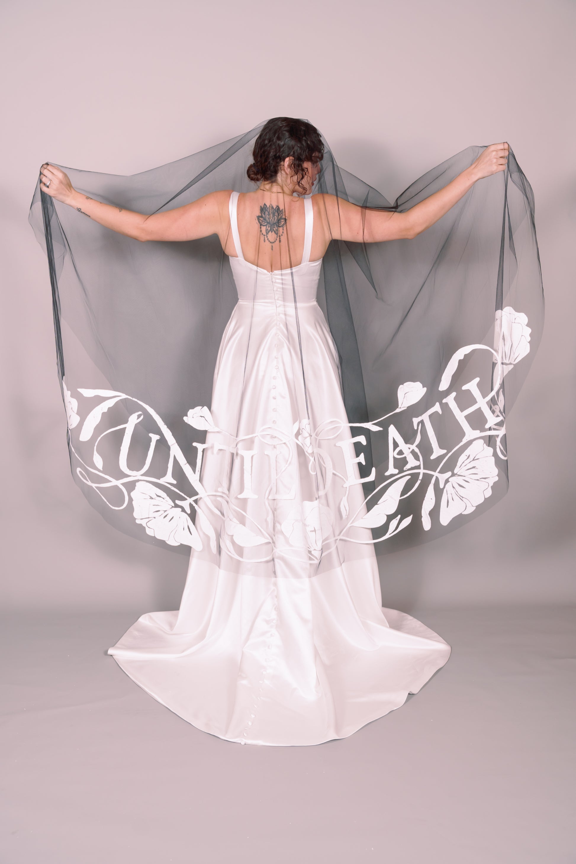 Woman in a white wedding dress with a long black painted veil that says Until Death against a plain background. Photography by Rachel Sakakeeny Model is Isabelle Paulino