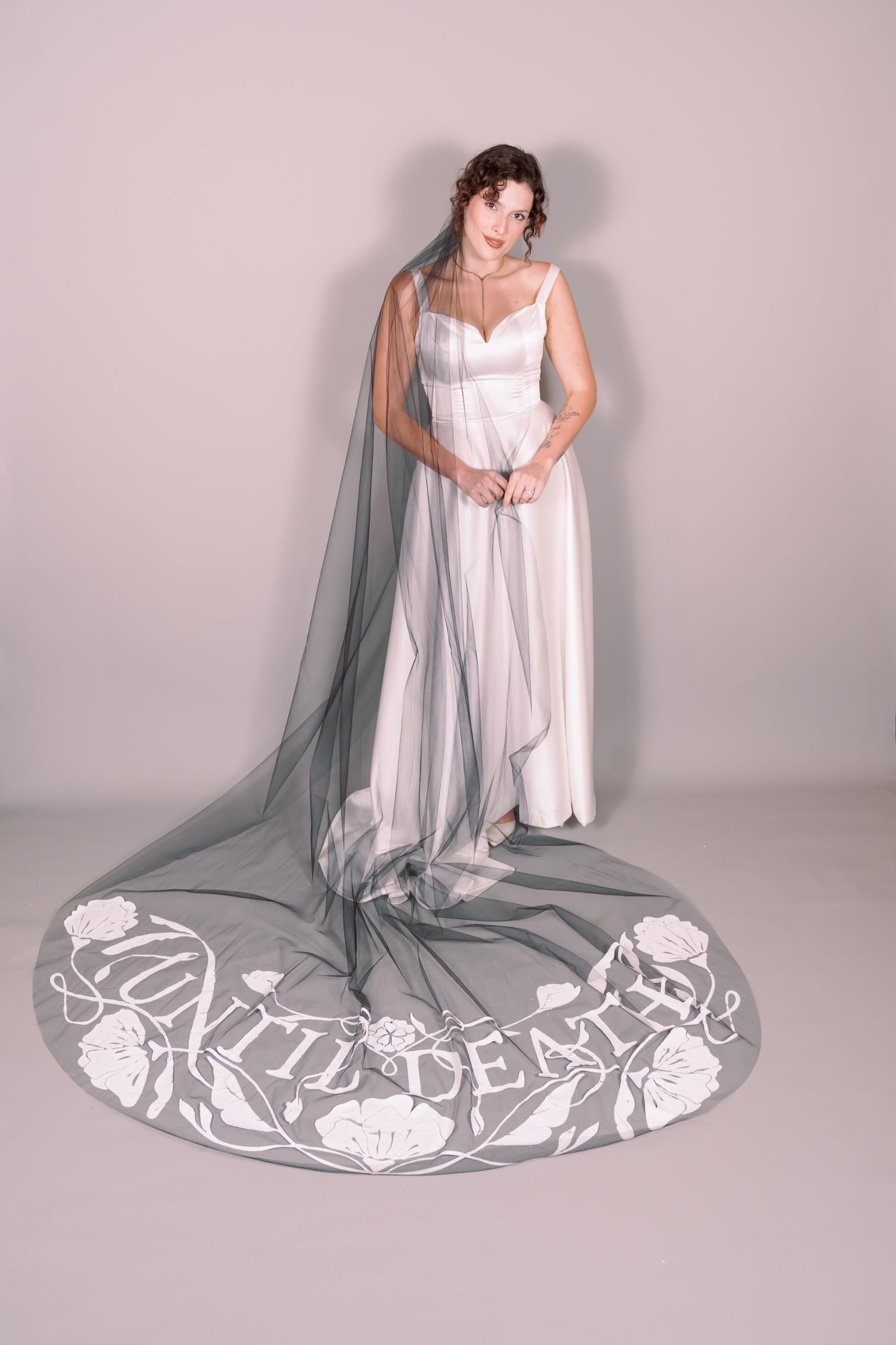 Woman in a white wedding dress with a long black painted veil that says Until Death against a plain background. Photography by Rachel Sakakeeny Model is Isabelle Paulino