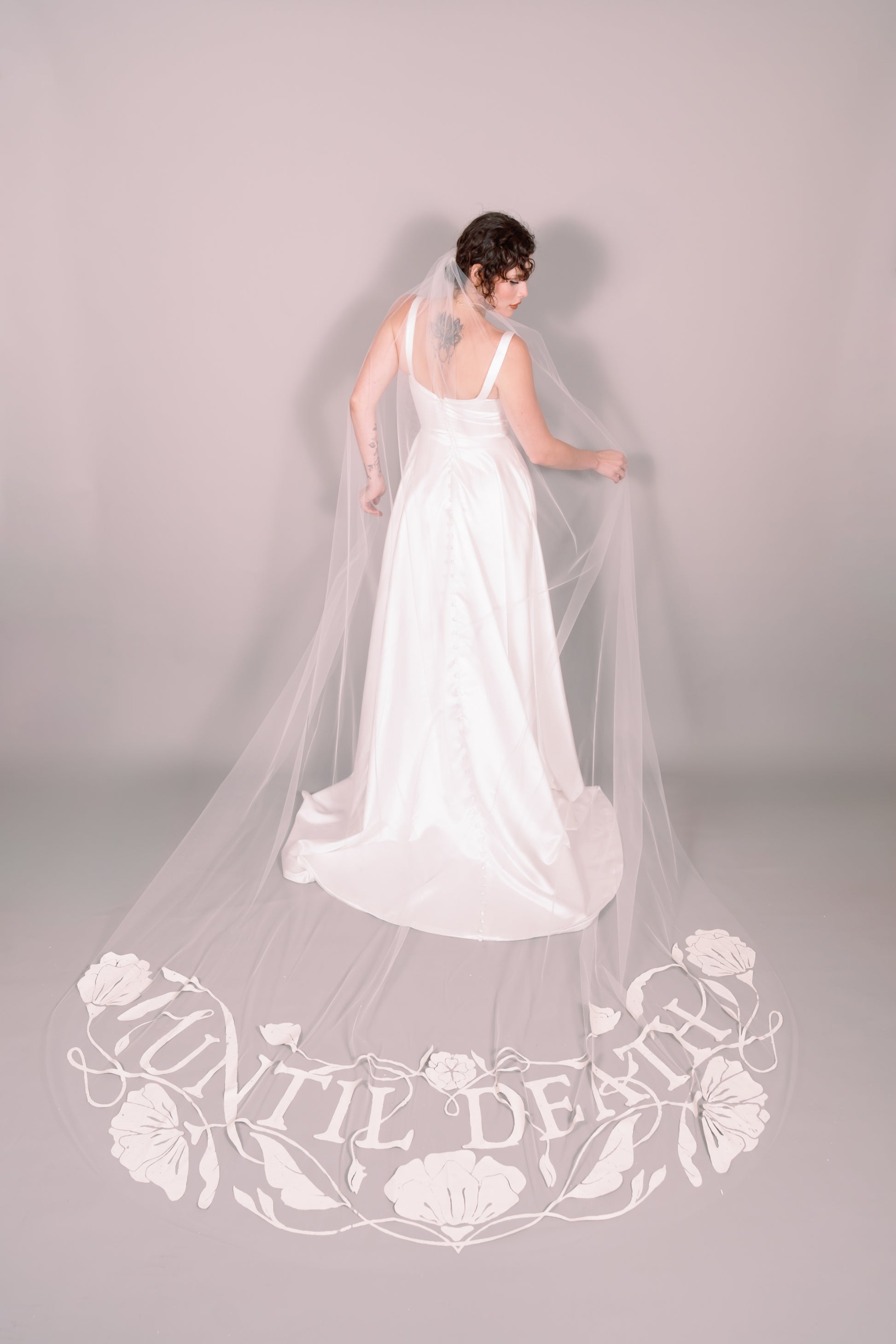 Woman in a white wedding dress with a long painted veil that says Until Death against a plain background. Photography by Rachel Sakakeeny Model is Isabelle Paulino