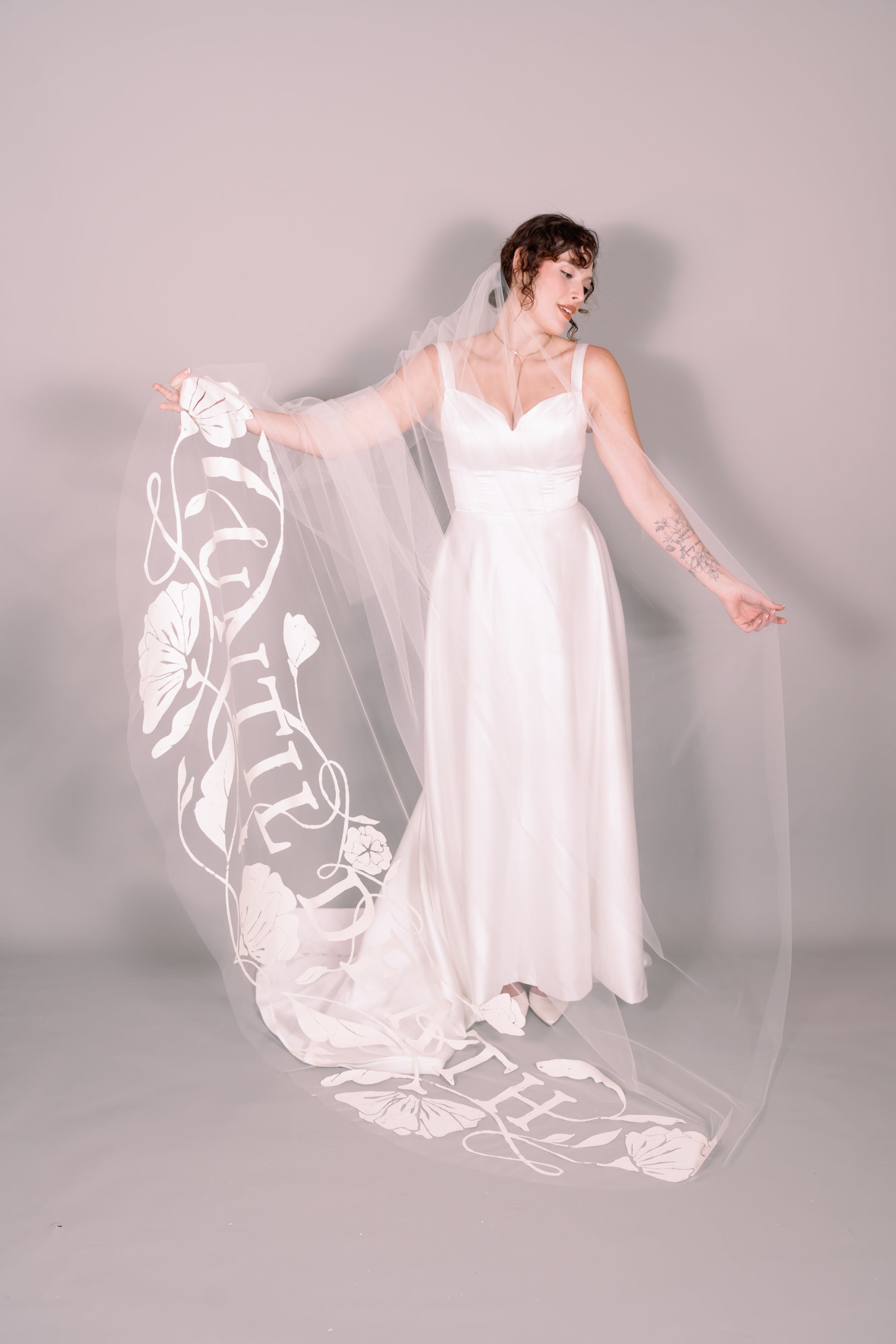 Woman in a white wedding dress with a long painted veil that says Until Death against a plain background. Photography by Rachel Sakakeeny Model is Isabelle Paulino
