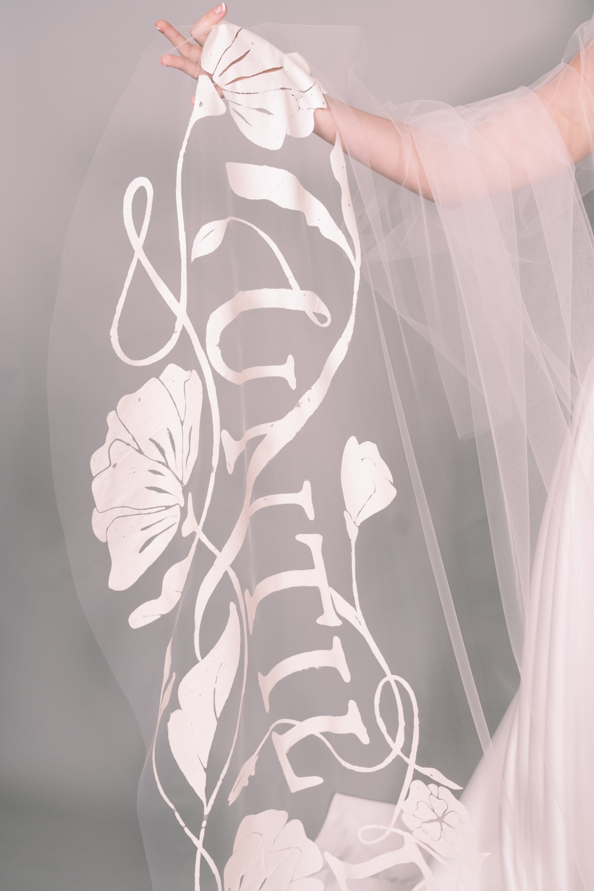 Veil with floral and leaf patterns, held by a hand against a neutral background. Painted veil says Until Death. Photography by Rachel Sakakeeny Model is Isabelle Paulino