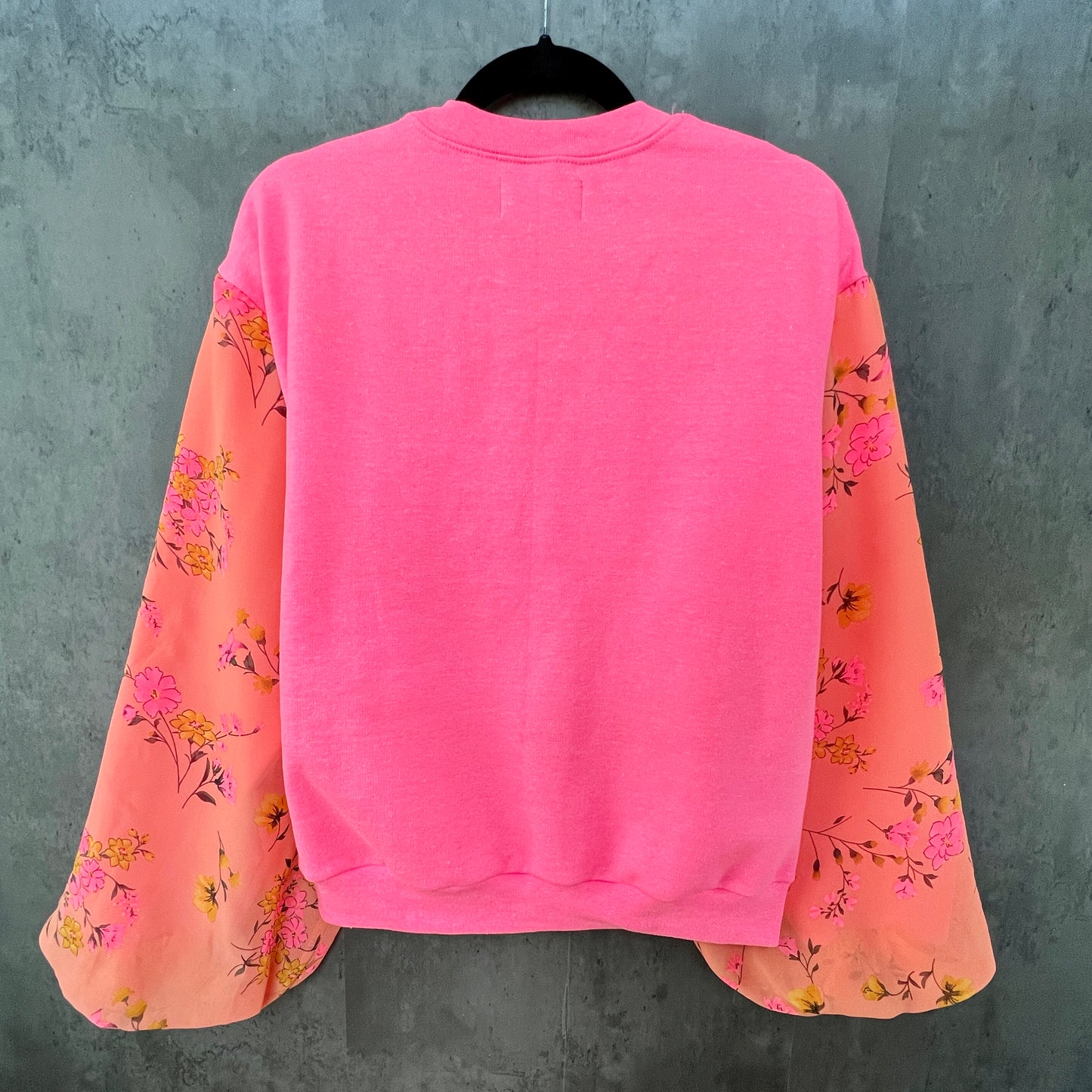 Back view of neon hot pink sweatshirt on hanger in front of cement gray background. Sweatshirt has puff sleeves with bright coral pink and marigold colorful floral.