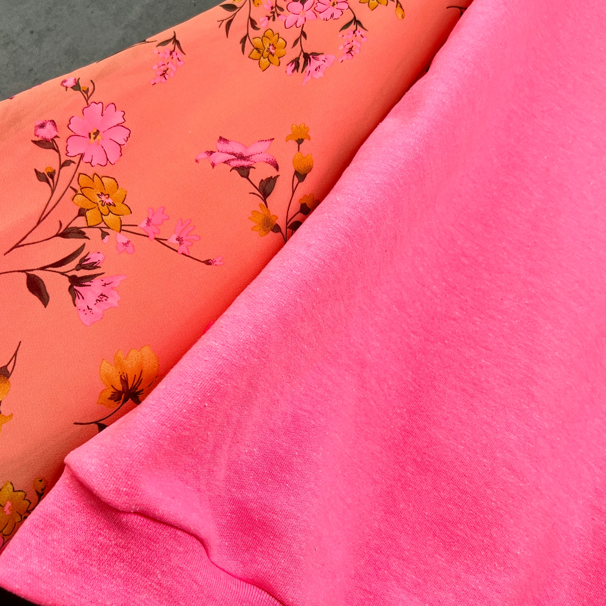 Detail view of neon hot pink sweatshirt on hanger in front of cement gray background. Sweatshirt has puff sleeves with bright coral pink and marigold colorful floral.