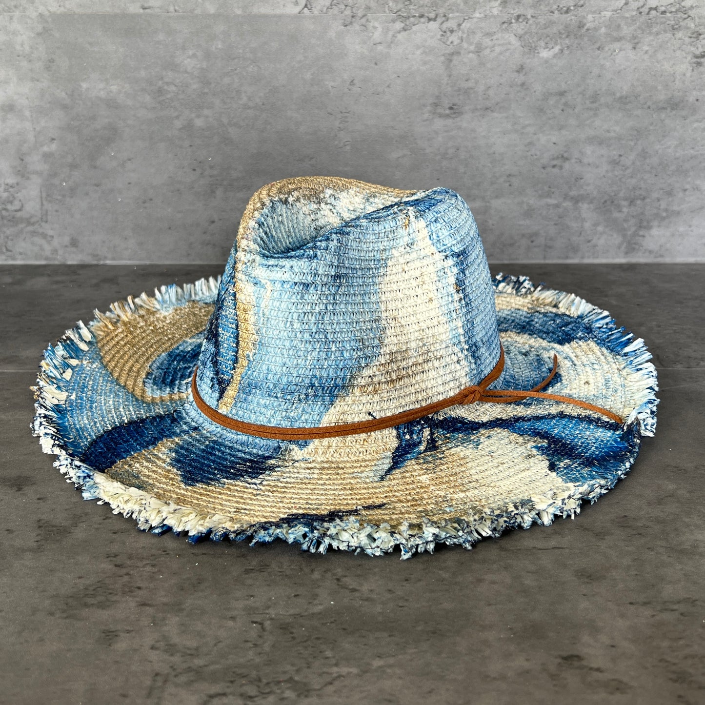 front view of cream white straw brim rancher hat with fringe has navy blue and gold paint marbling  and brown leather faux suede cord. sun hat.