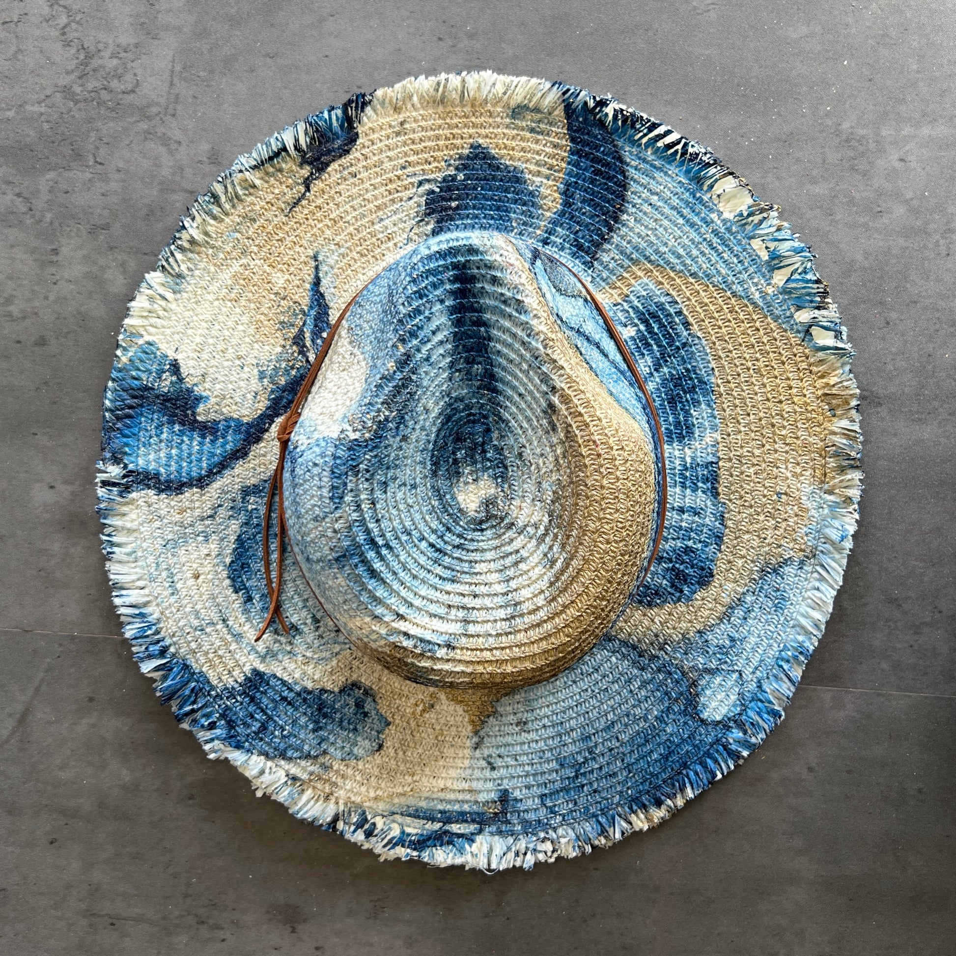 top view of cream white straw brim rancher hat with fringe has navy blue and gold paint marbling  and brown leather faux suede cord. sun hat.