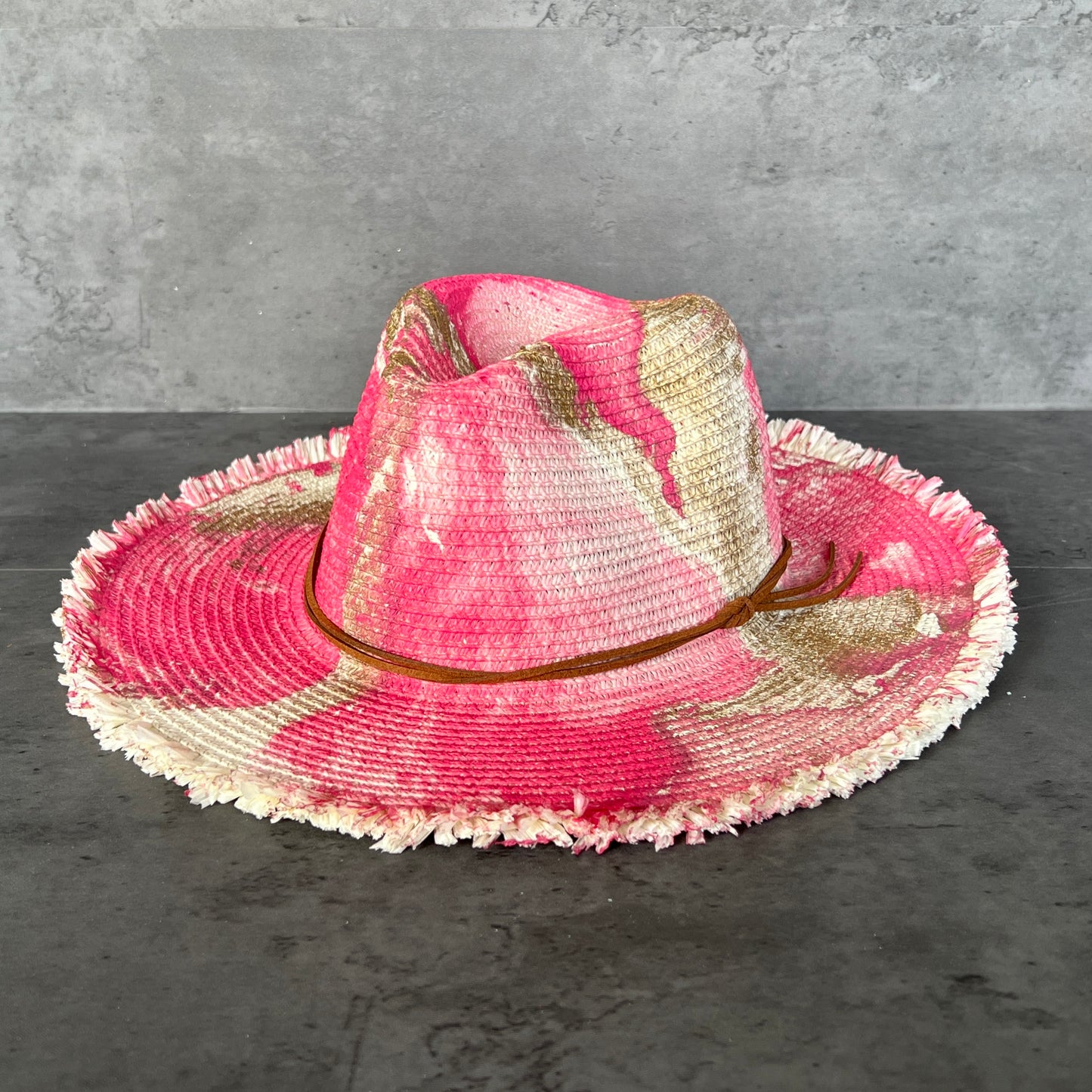 front view of cream white straw brim rancher hat with fringe has hot pink fuchsia and gold paint marbling  and brown leather faux suede cord. sun hat.
