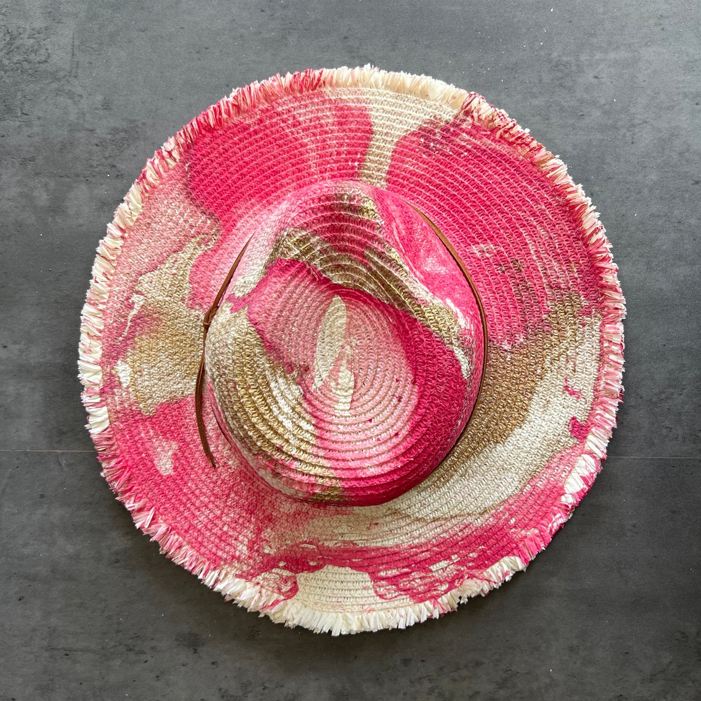 top view of cream white straw brim rancher hat with fringe has hot pink fuchsia and gold paint marbling  and brown leather faux suede cord. sun hat.