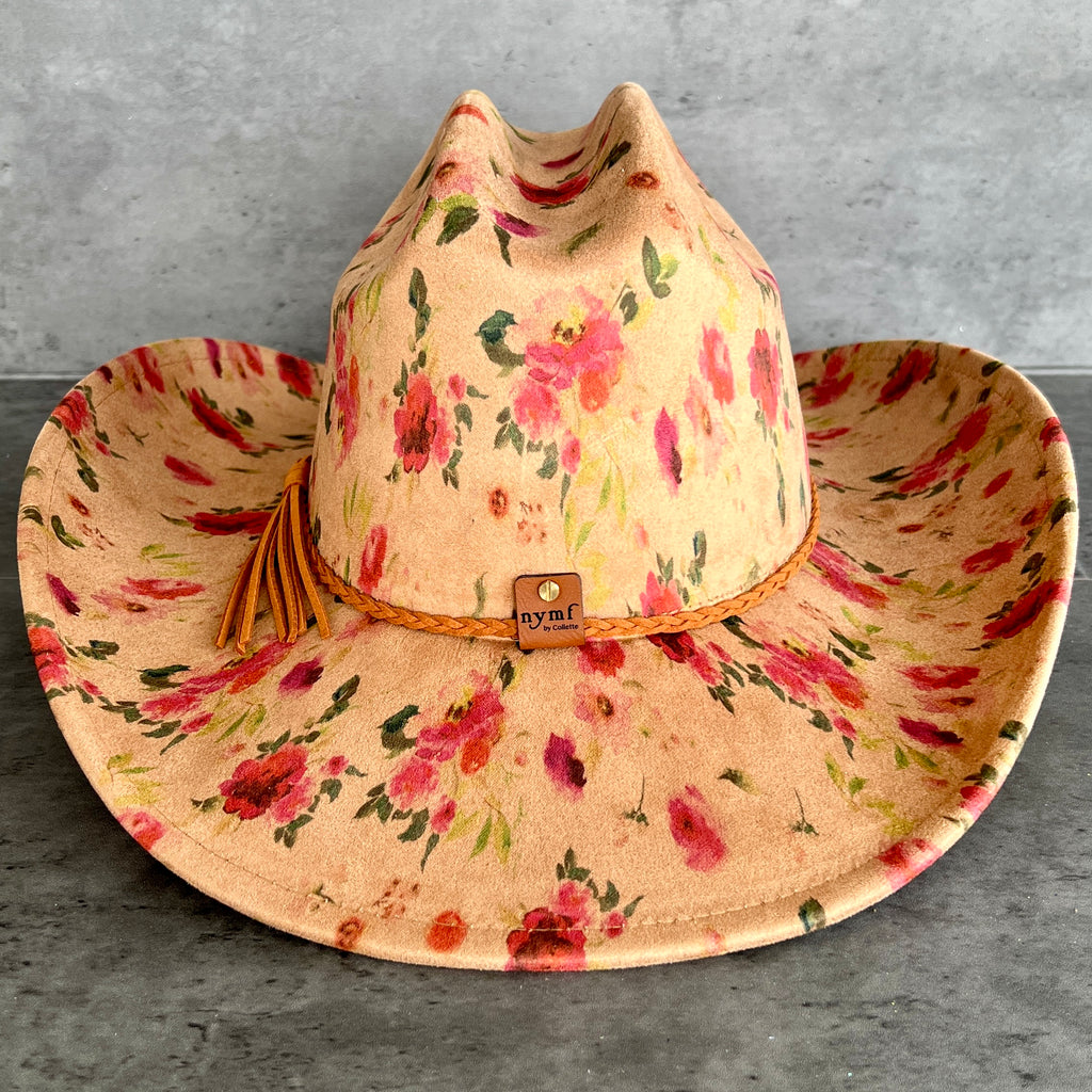 A wide-brimmed cowboy hat with a tan base and a pink, red, and green floral print, featuring a braided tan band with a small 'nymf' logo patch and a tassel.