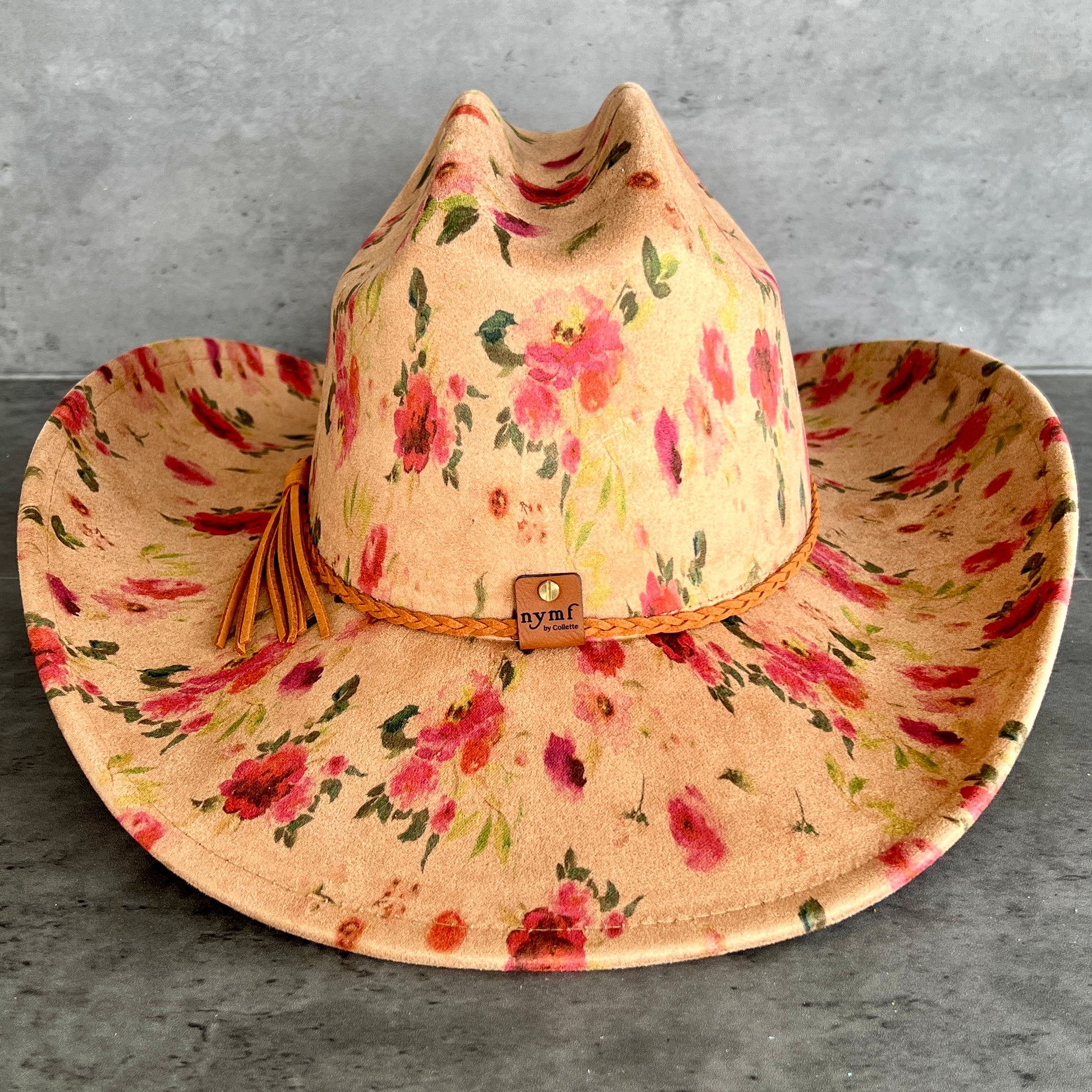 A wide-brimmed cowboy hat with a tan base and a pink, red, and green floral print, featuring a braided tan band with a small 'nymf' logo patch and a tassel.