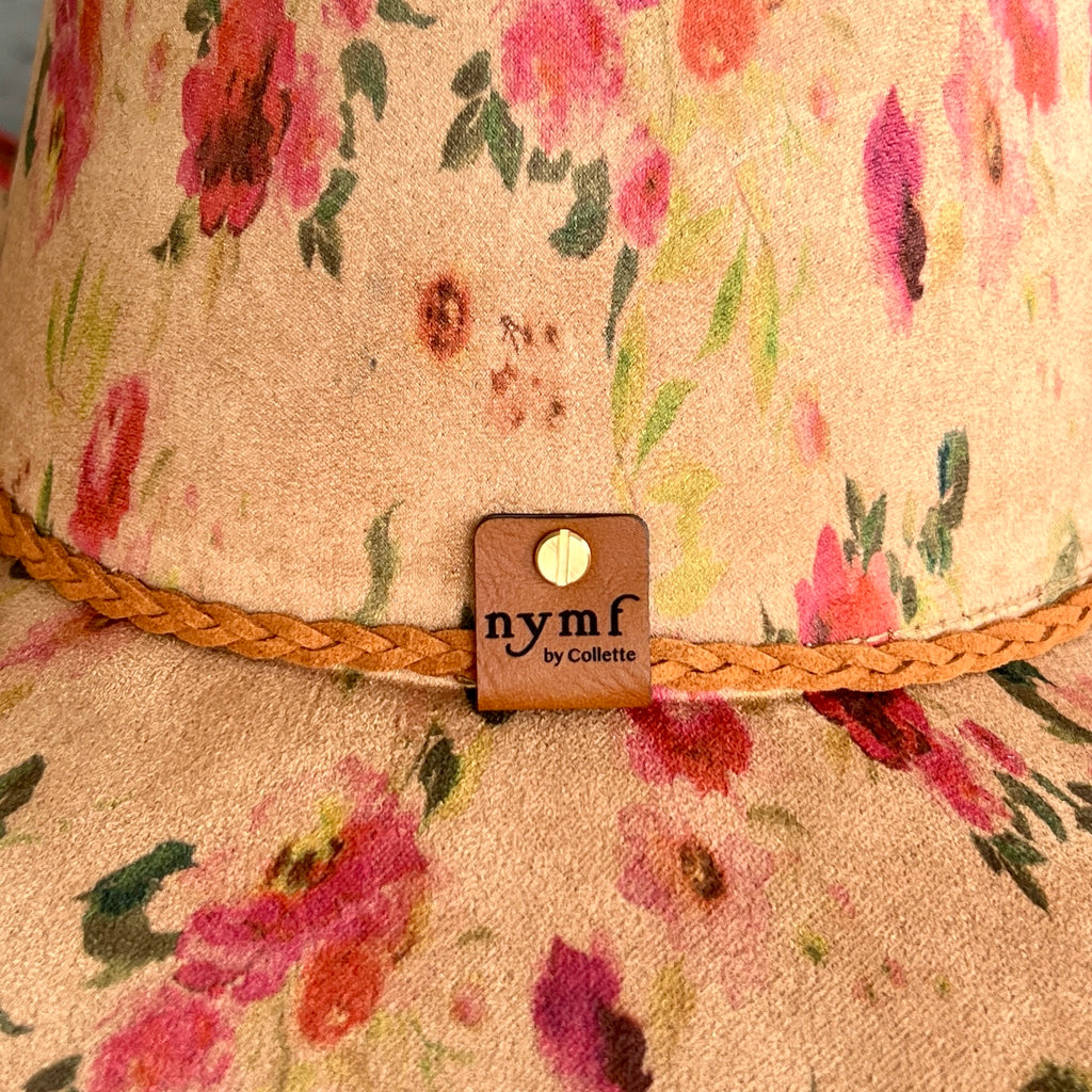 Close-up of a floral cowboy hat with a tan base, pink and red flowers, green leaves, and a braided tan band. A small brand tag reading 'nymf by Collette' is attached at the front.