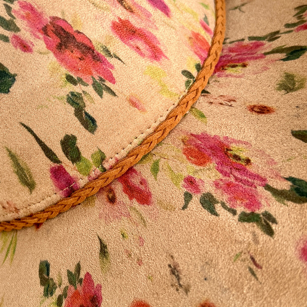 Close-up of a floral hat with pink and red flowers, green leaves, and a braided tan trim on a tan fabric background.