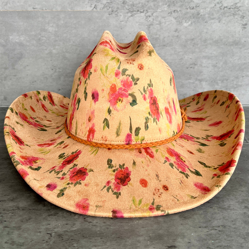 A western-style cowboy hat with a tan base and a pink, red, and green floral pattern, featuring a braided tan band around the crown, displayed on a gray textured surface.