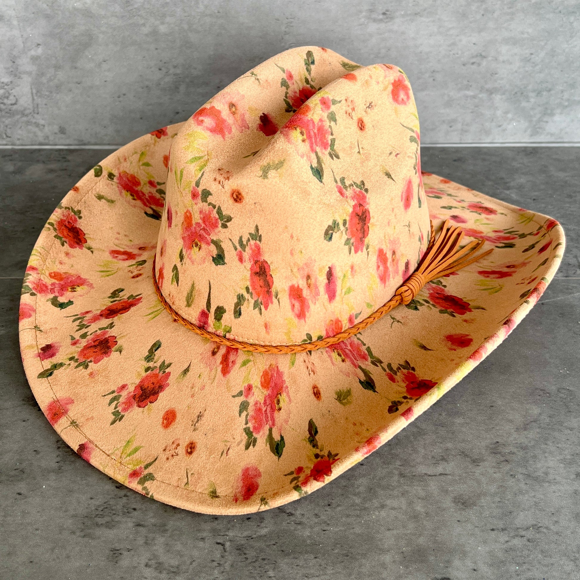 A wide-brimmed cowboy hat with a tan base and a red and green floral print, resting on a gray textured surface.