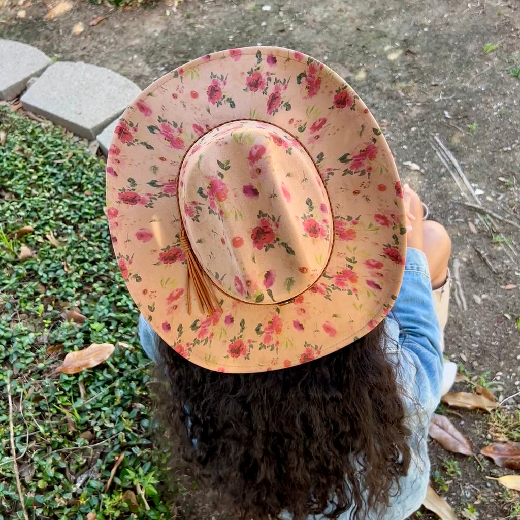 A cowboy hat with a pink and red floral print on a beige base, worn by a person sitting outdoors on a dirt path with green plants on the left.