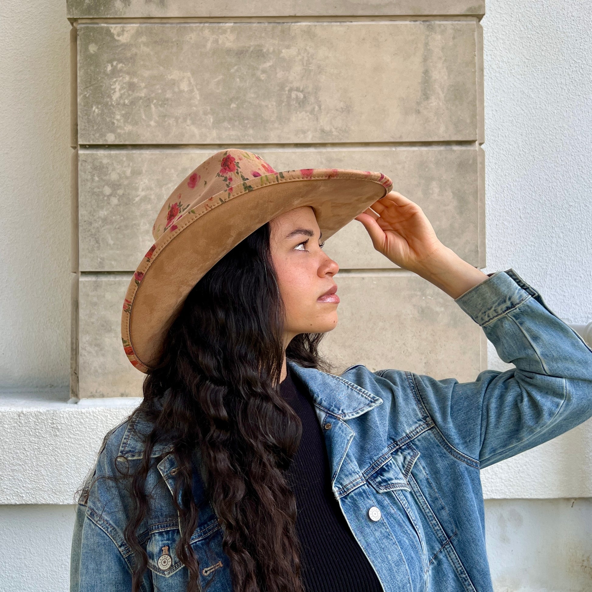 A woman wearing a tan cowboy hat with a red floral pattern on the crown and a blue denim jacket, posing in front of a textured stone wall.