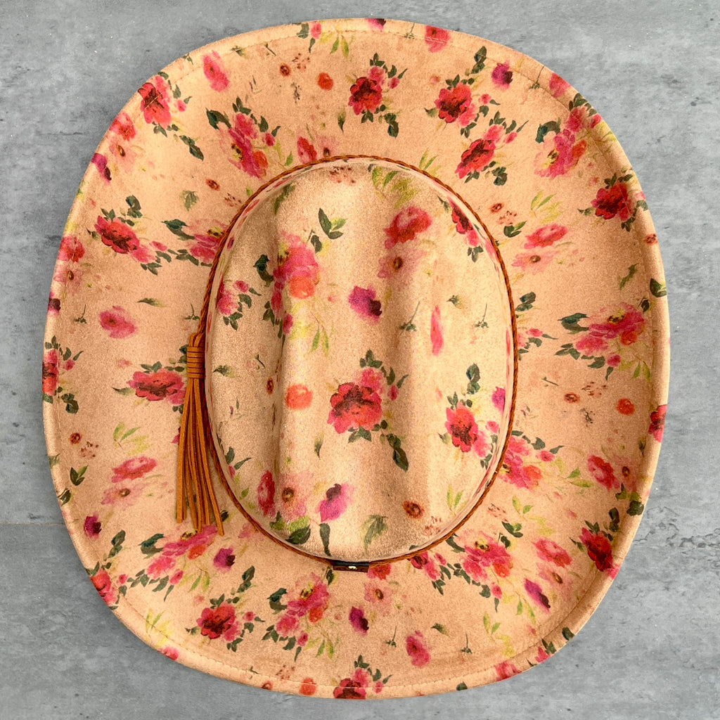 A wide-brimmed cowboy hat with a tan base and a vibrant pink, red, and green floral print and a brown leather tassel on the side, laid flat on a gray textured surface.