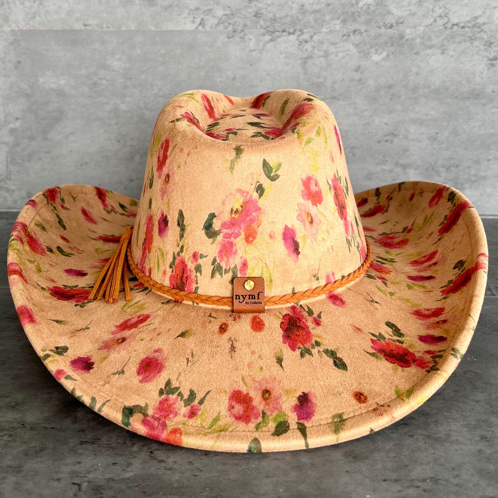 A floral cowboy hat with a tan base, pink and red flowers, and green leaves, featuring a braided tan band with a small 'nymf' logo patch.