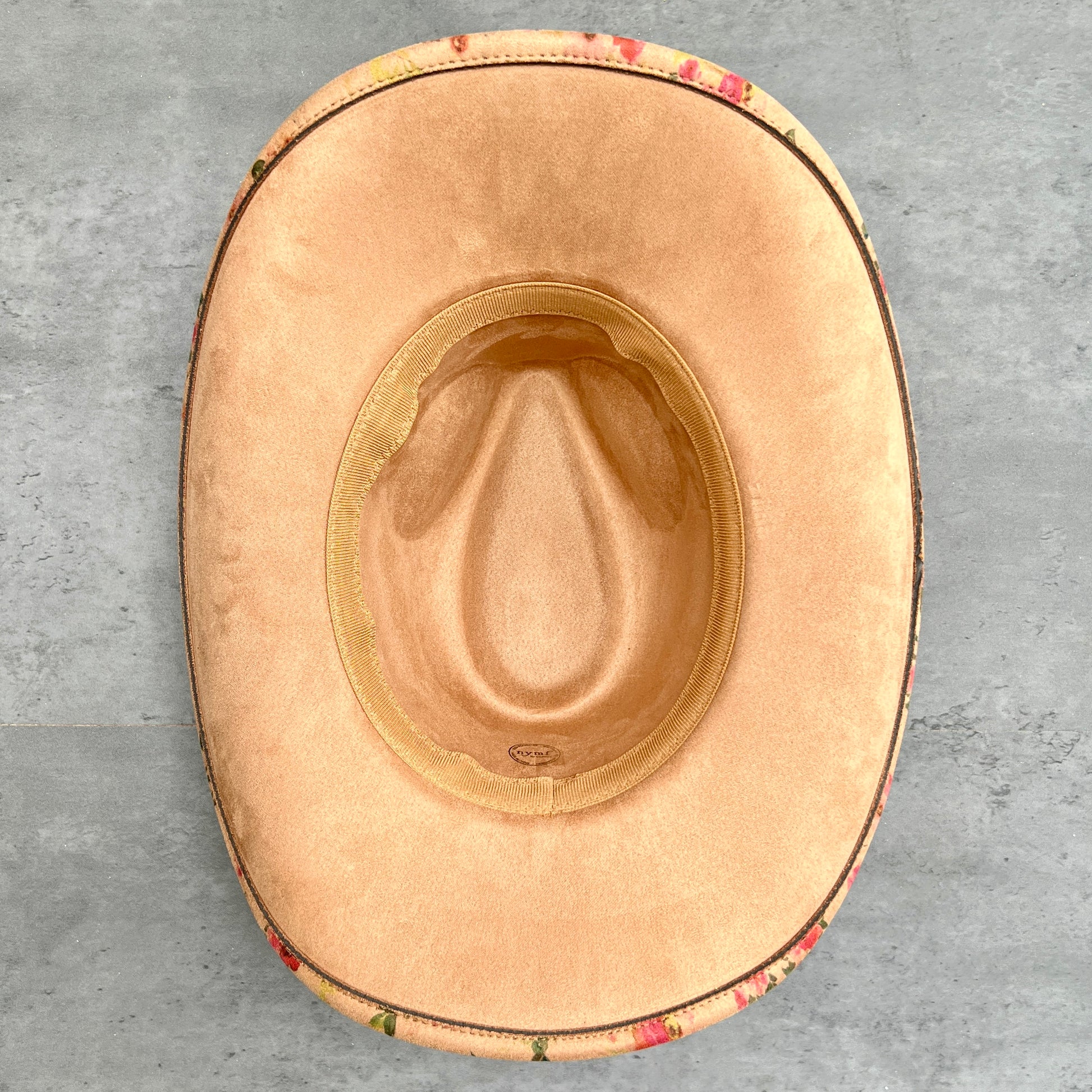 Base view of a tan cowboy hat with a floral patterned brim, set against a gray concrete background.