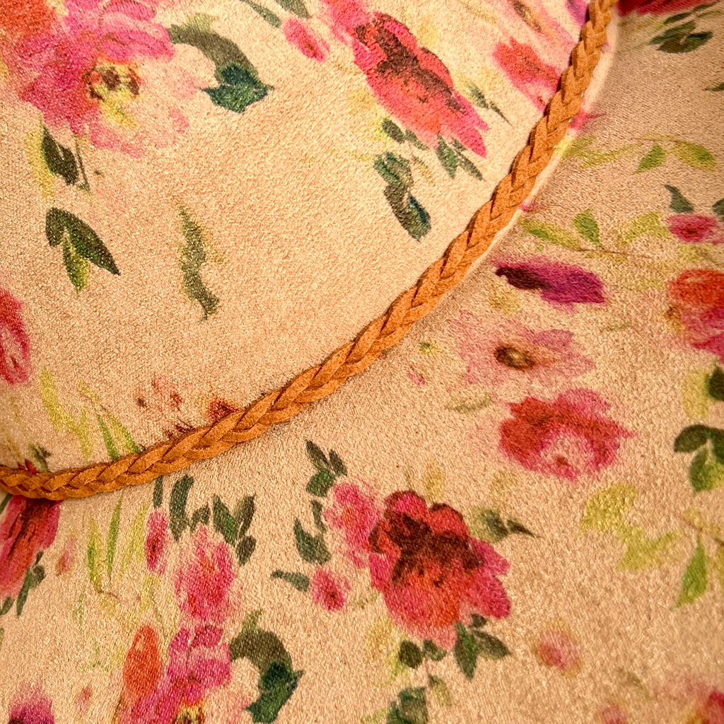 Close-up of a floral hat with pink and red flowers, green leaves, and a braided tan rope trim on a tan fabric. 
