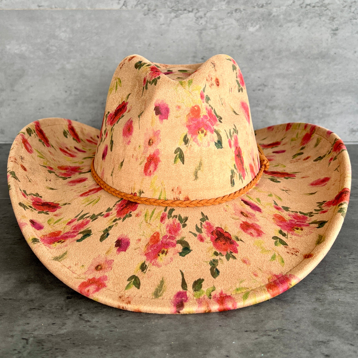 A wide-brimmed cowboy hat with a tan base and a pink, red, and green floral pattern, featuring a braided tan band around the crown, set against a gray textured background.