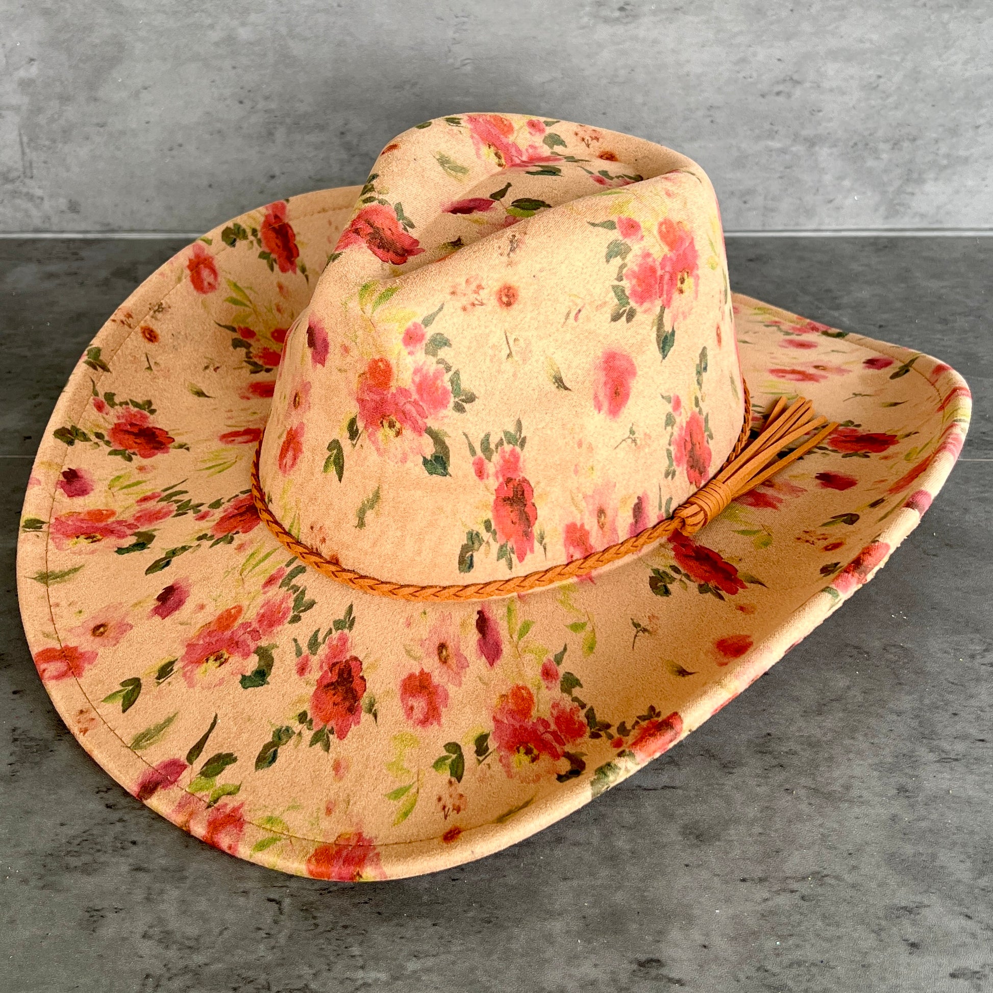 A wide-brimmed cowboy hat with a tan base and a pink and green floral print, featuring a braided tan band and tassel, displayed on a gray stone surface.