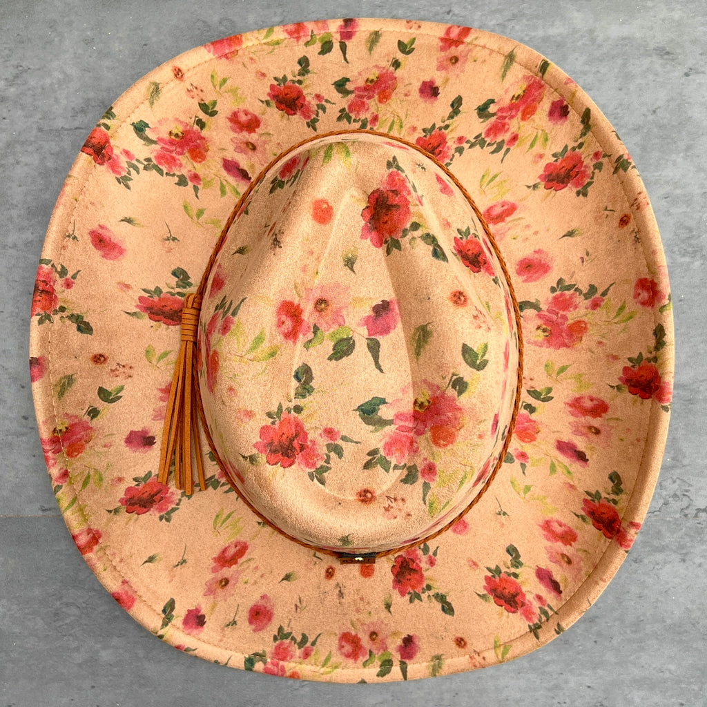 A wide-brimmed cowboy hat with a tan floral print, featuring red and pink flowers with green leaves, a brown leather trim, and a matching straw tassel on the side, photographed from a top-down view on a gray textured background.