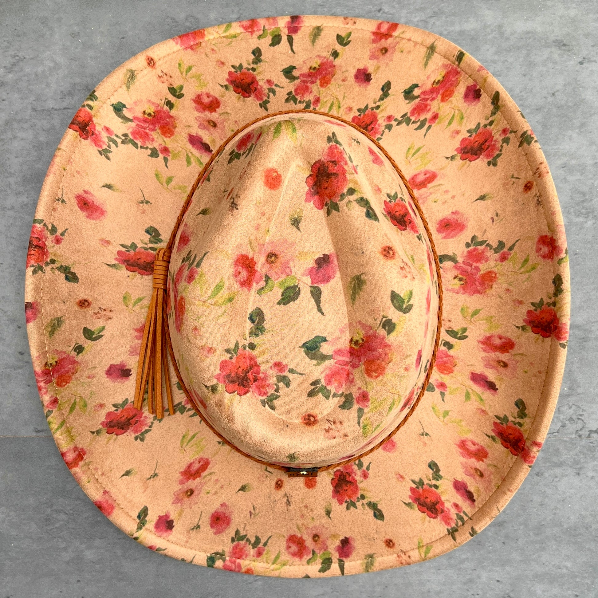 A wide-brimmed cowboy hat with a tan floral print, featuring red and pink flowers with green leaves, a brown leather trim, and a matching straw tassel on the side, photographed from a top-down view on a gray textured background.