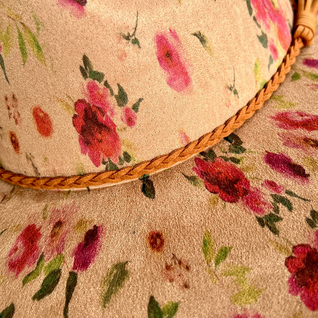 Close-up of a floral suede hat, featuring a tan base with pink, red, and green watercolor-style flowers and a braided tan rope trim.