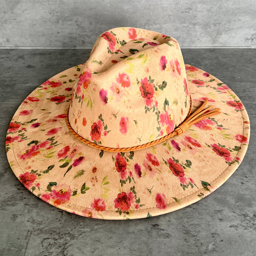 A wide-brimmed sun hat with a beige floral print, featuring pink and red flowers with green leaves, a braided tan band, and a small tassel, displayed on a gray concrete surface.