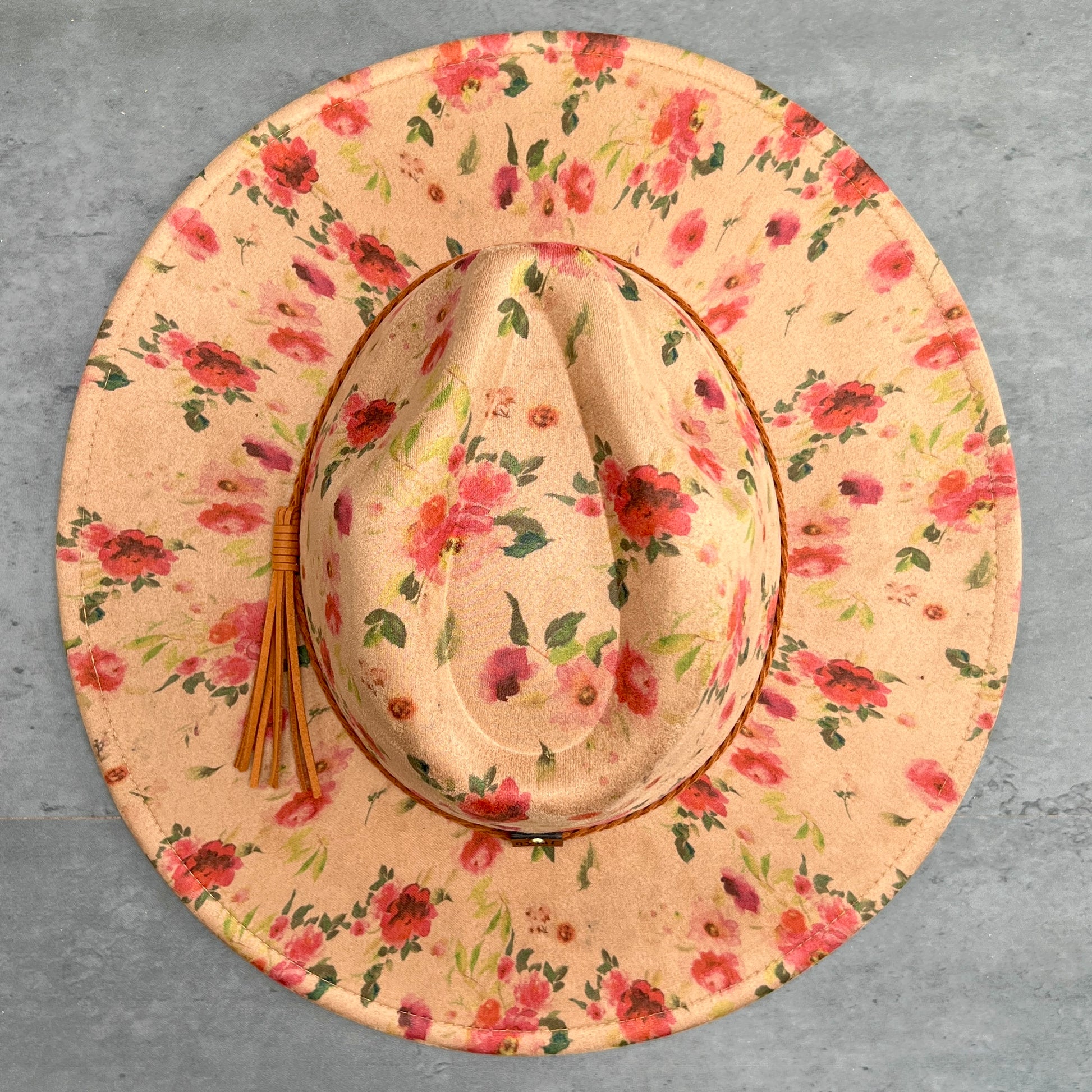A wide-brimmed cowboy hat with a floral print, featuring a tan leather tassel on the side, photographed from directly above on a textured gray background.
