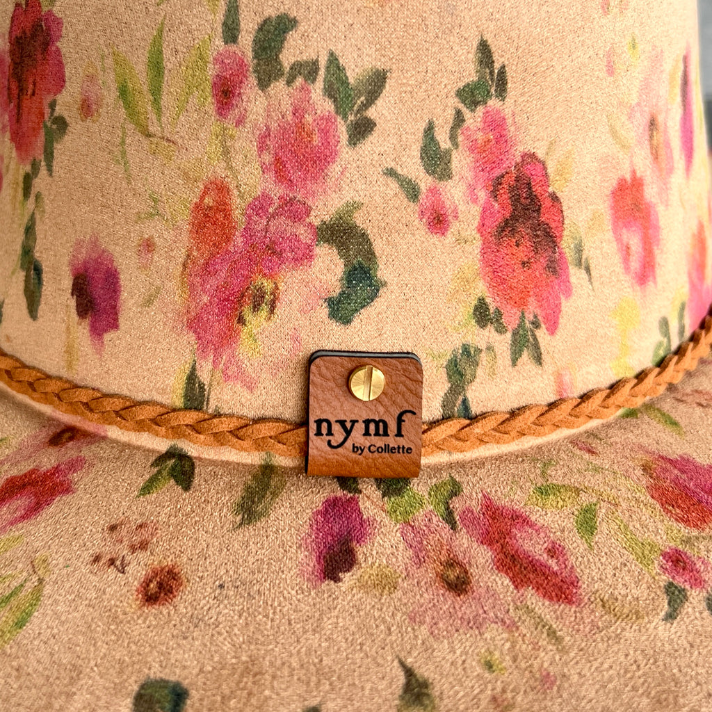 Close-up of a floral hat with pink and red flowers on a beige textured fabric, featuring a braided tan rope band and a small brand tag that reads 'nymf by Collette'.