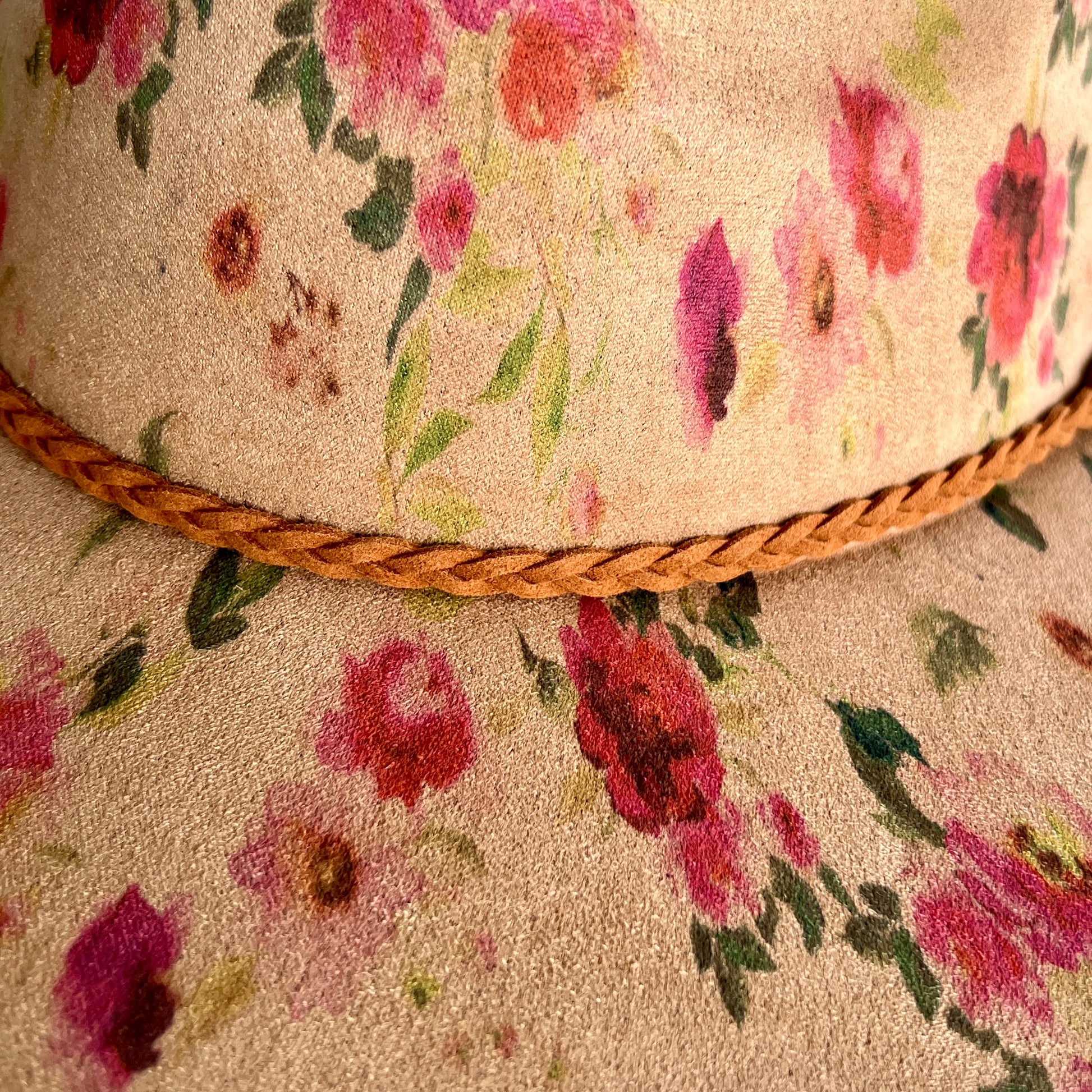 Close-up of a floral hat with a tan base, pink and red watercolor-style flowers, green leaves, and a braided tan rope band around the crown.
