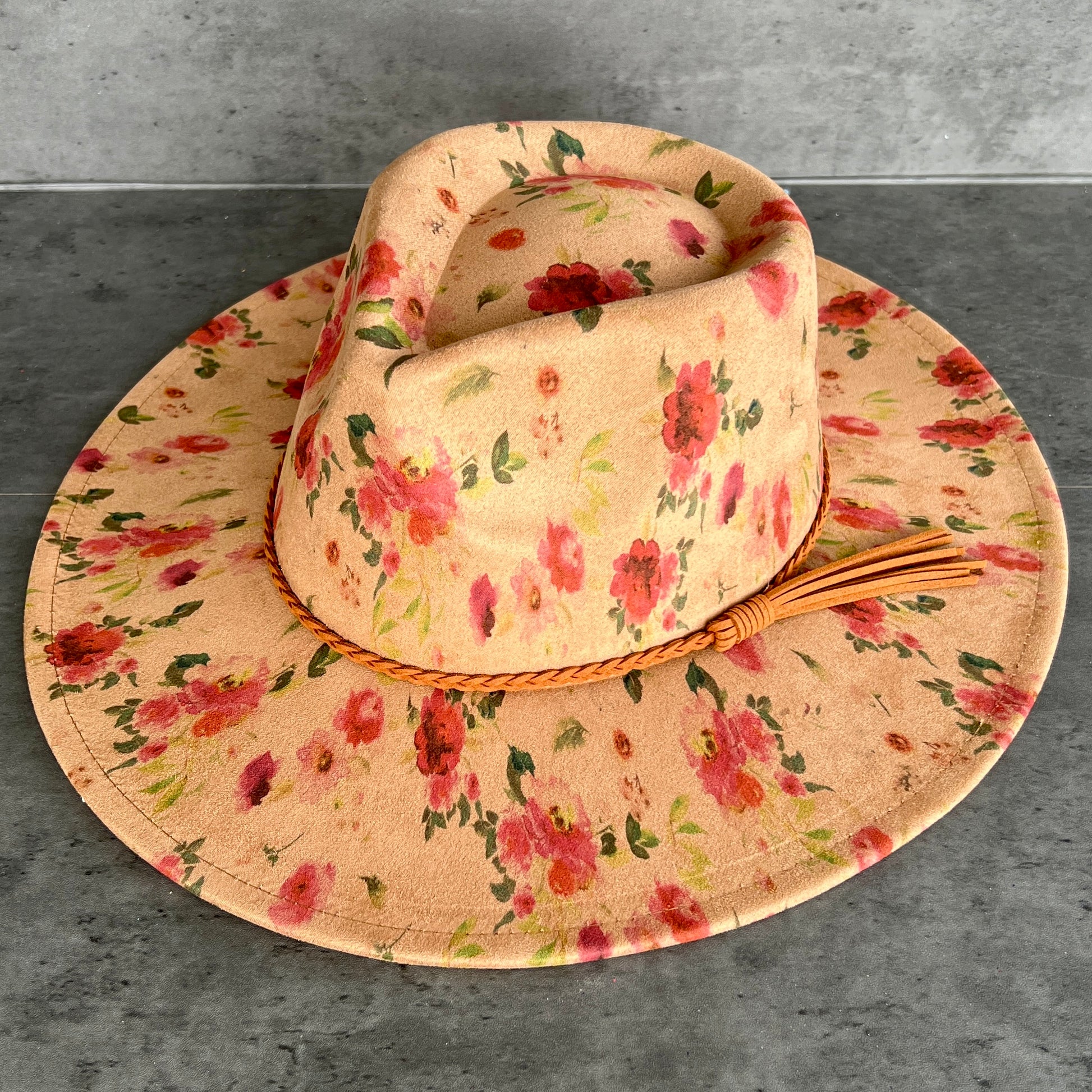 A wide-brimmed sun hat with a tan floral print, featuring red and pink flowers with green leaves, a braided tan band, and a tassel, displayed on a gray textured surface.