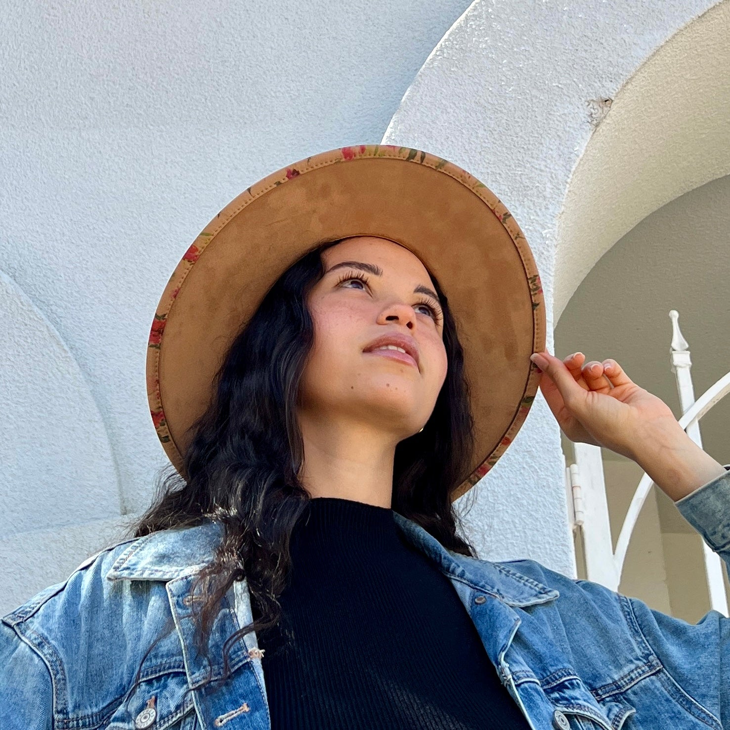 A woman wearing a wide-brimmed tan hat with a colorful patterned inner band, a black ribbed top, and a light blue denim jacket, posing outdoors against a white stucco wall with arches.