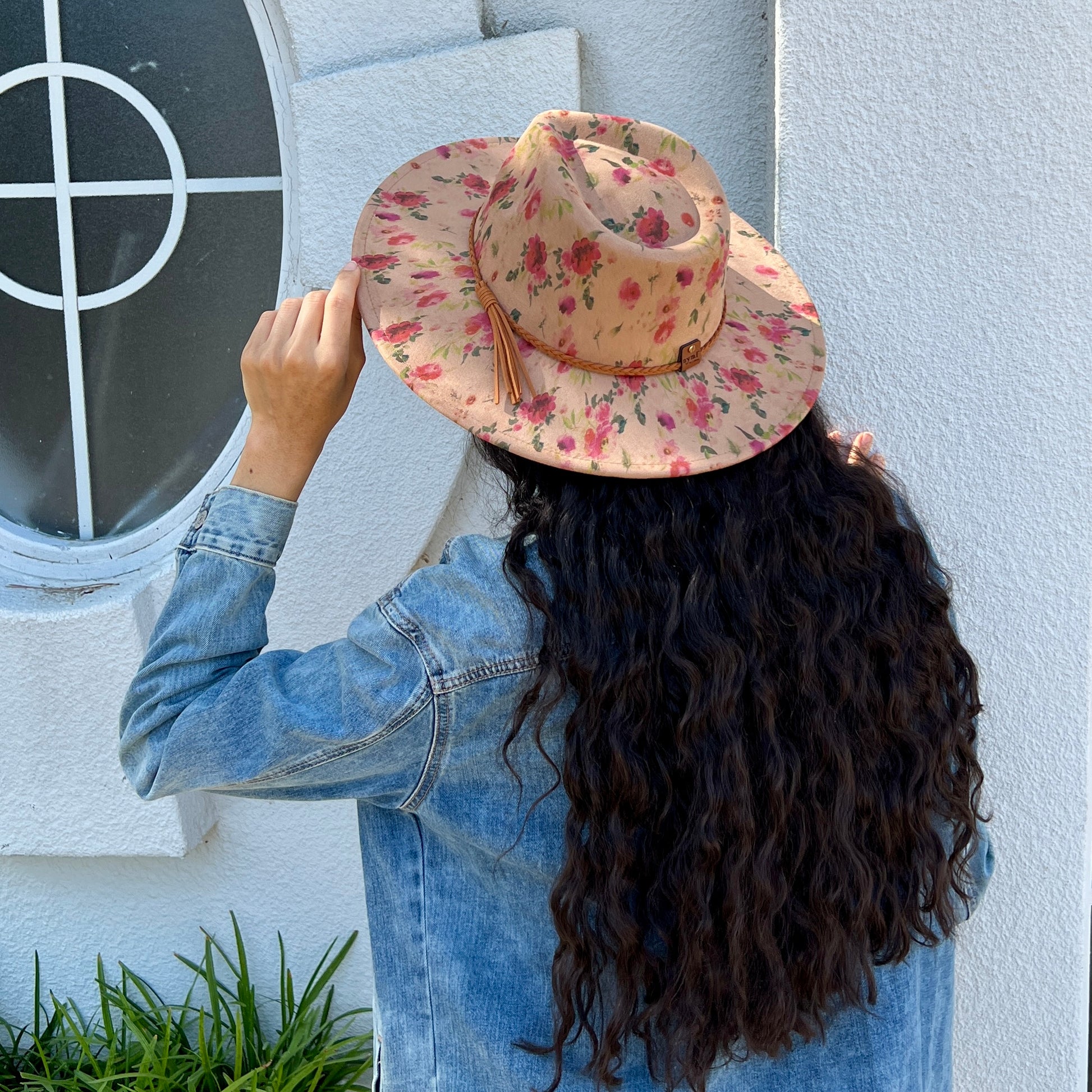 A person viewed from behind wearing a wide-brimmed tan hat with a pink and red floral pattern, a light blue denim jacket, and long dark curly hair, standing outdoors against a white stucco wall near a window.