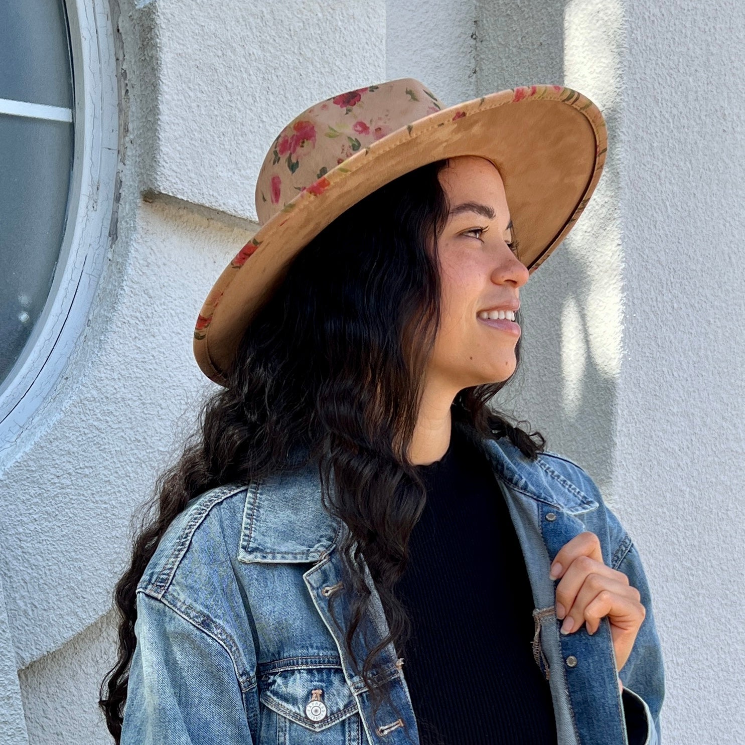A woman wearing a wide-brimmed suede hat with a pink floral pattern and a blue denim jacket, standing against a textured white stucco wall.