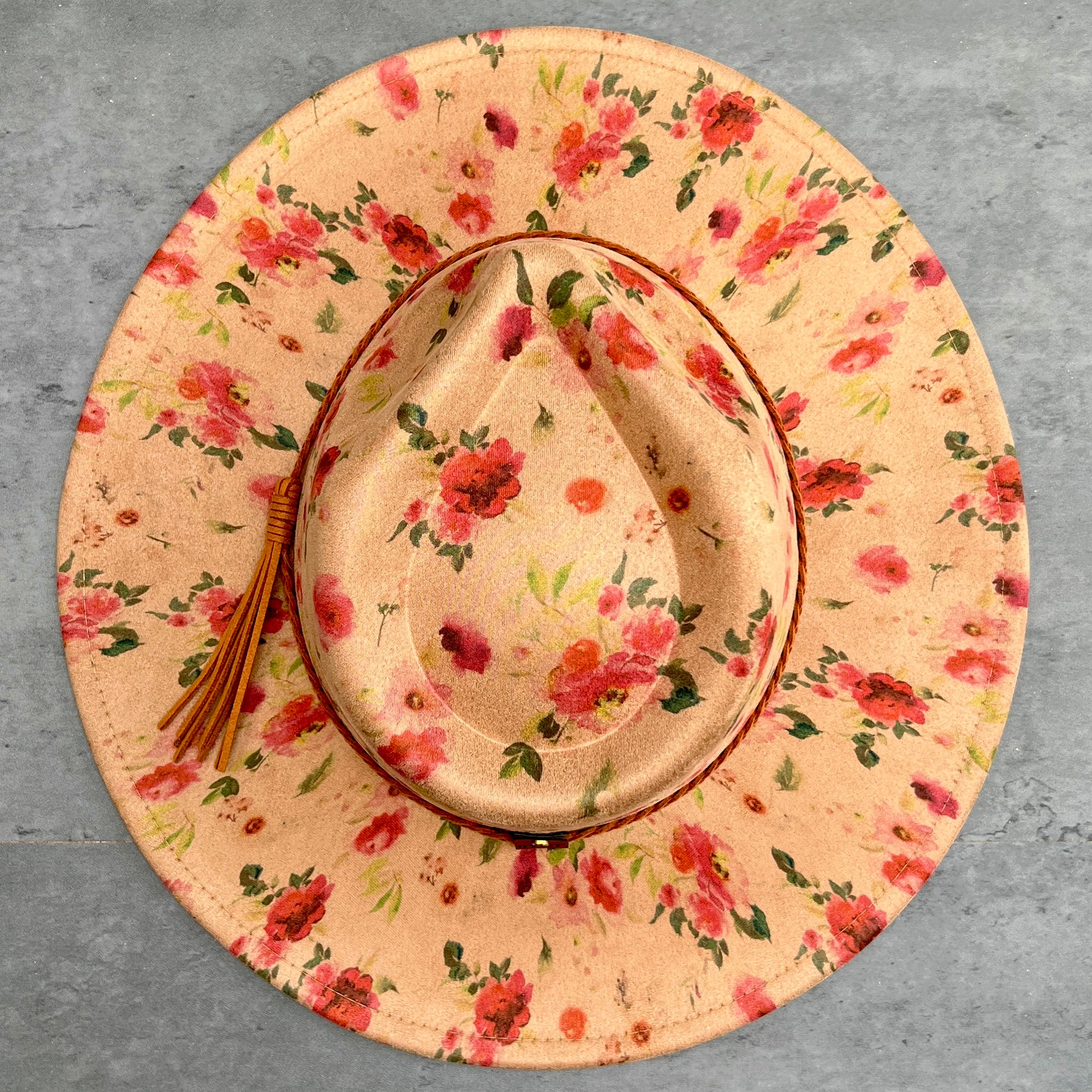 A wide-brimmed cowboy hat with a tan floral print, featuring red and pink flowers with green leaves, a brown leather-look band, and a brown tassel on the side, photographed from a top-down view on a gray textured background.
