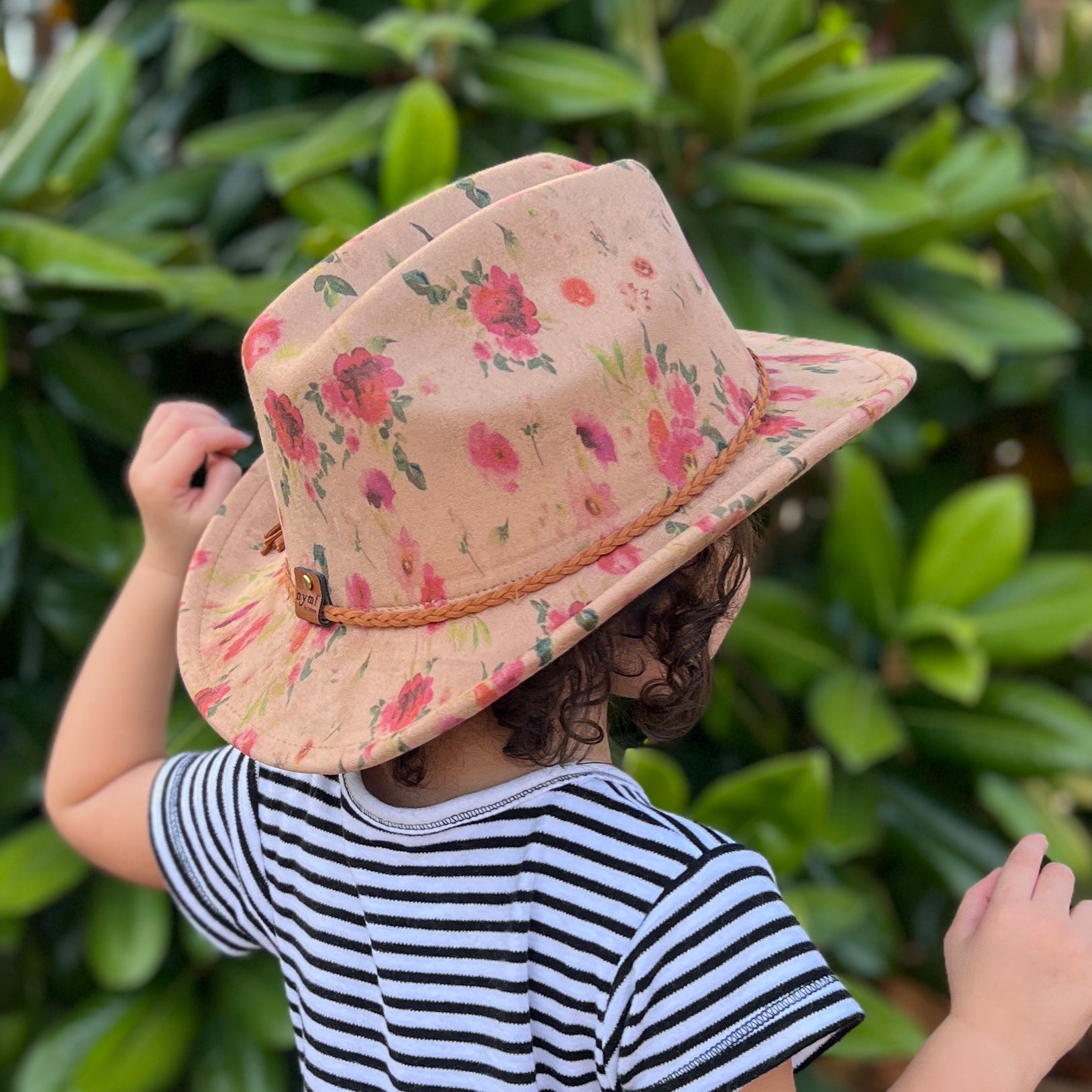 A child seen from behind wearing a small cowboy hat with a pink and red floral print on a beige background, paired with a black and white striped shirt, set against a background of green foliage.