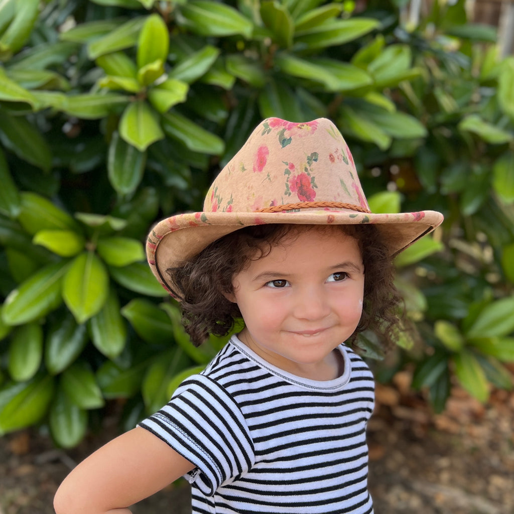 A young child wearing a tan floral cowboy hat and a black and white striped shirt, standing outdoors in front of green foliage.