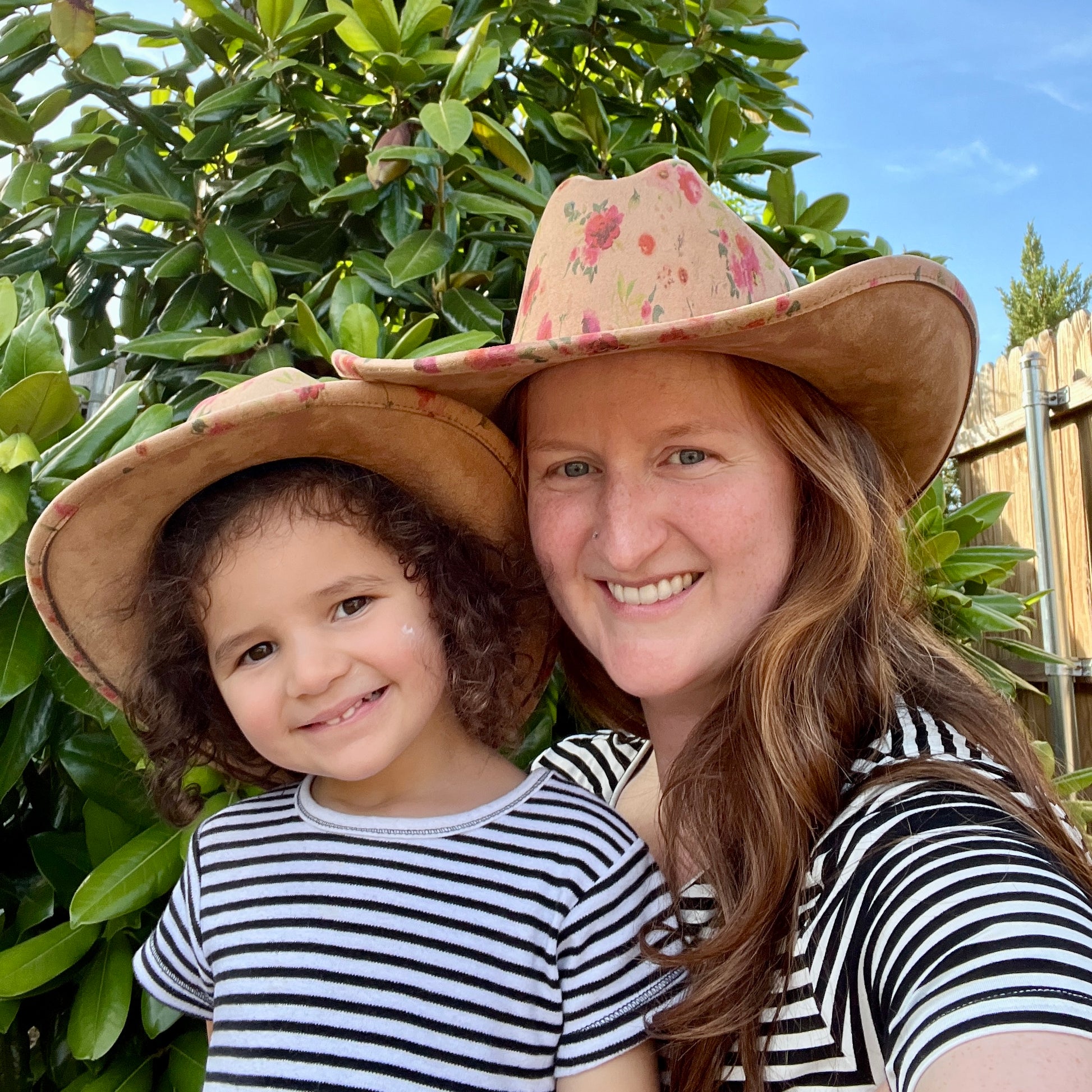A woman and a young girl wearing matching floral cowboy hats and striped shirts, smiling outdoors in front of green garden foliage.