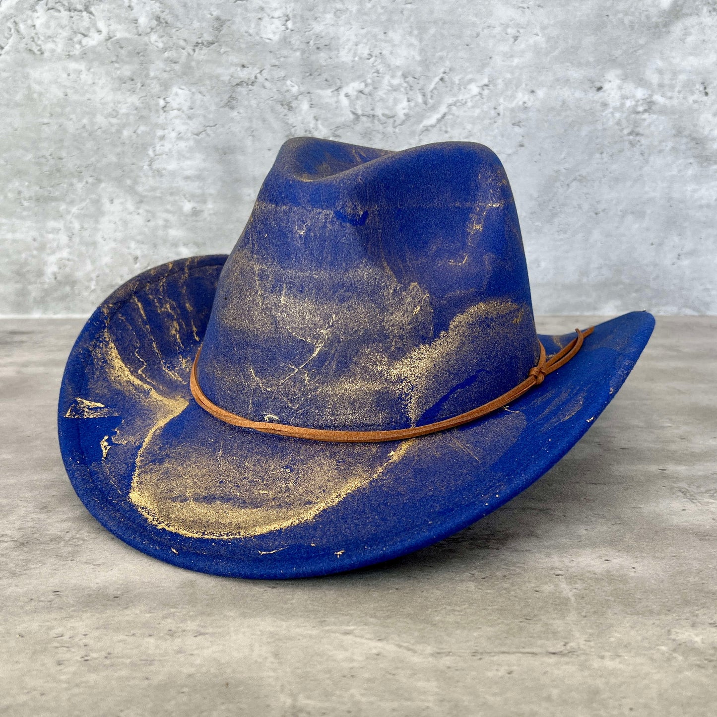 Royal cobalt blue western cowboy hat painted with gold marbling. Has light brown faux suede cord hat band. 