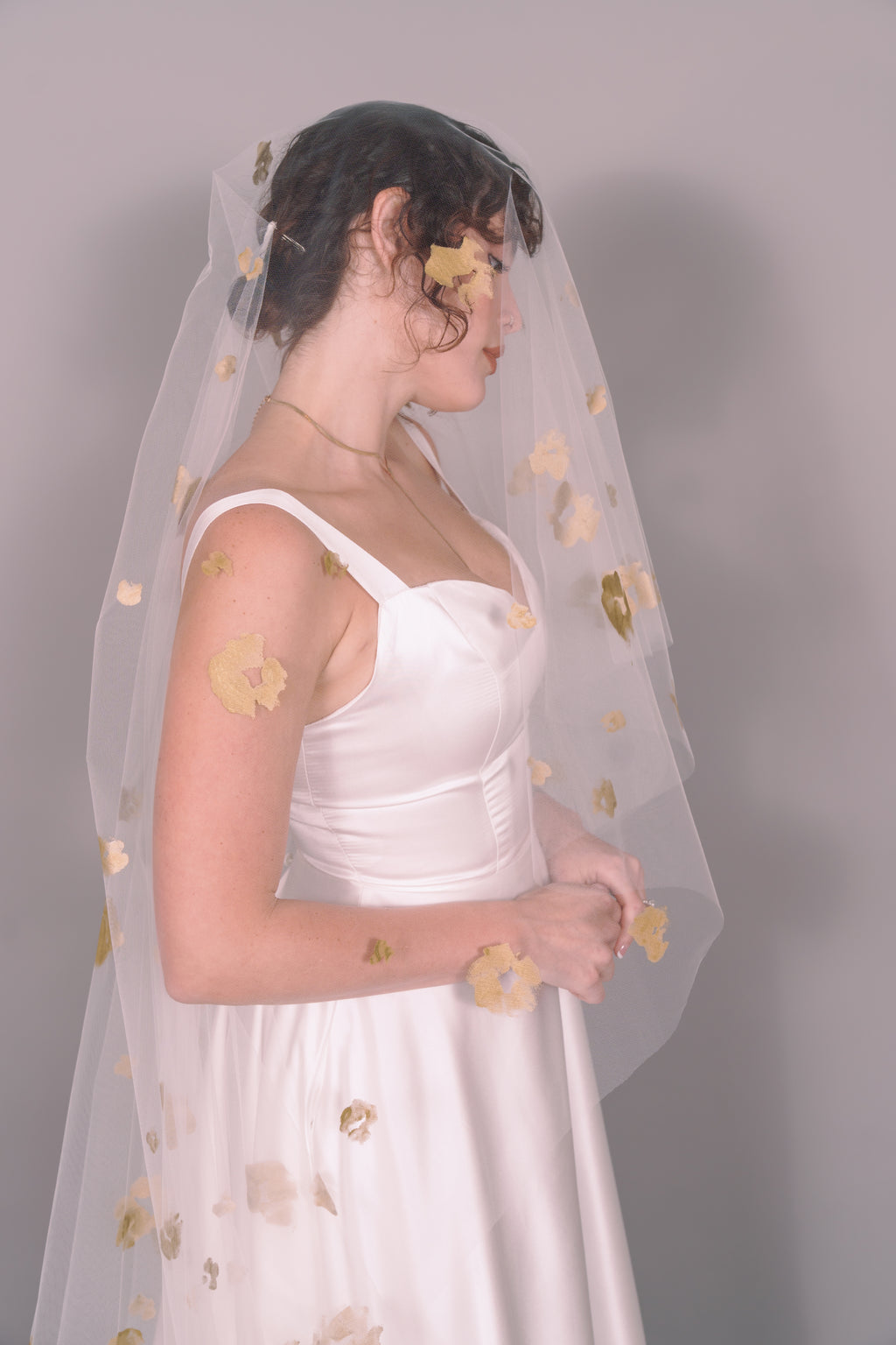 Woman wearing a wedding dress and ivory veil with painted gold floral petals on a plain background. Photography by Rachel Sakakeeny Model is Isabelle Paulino