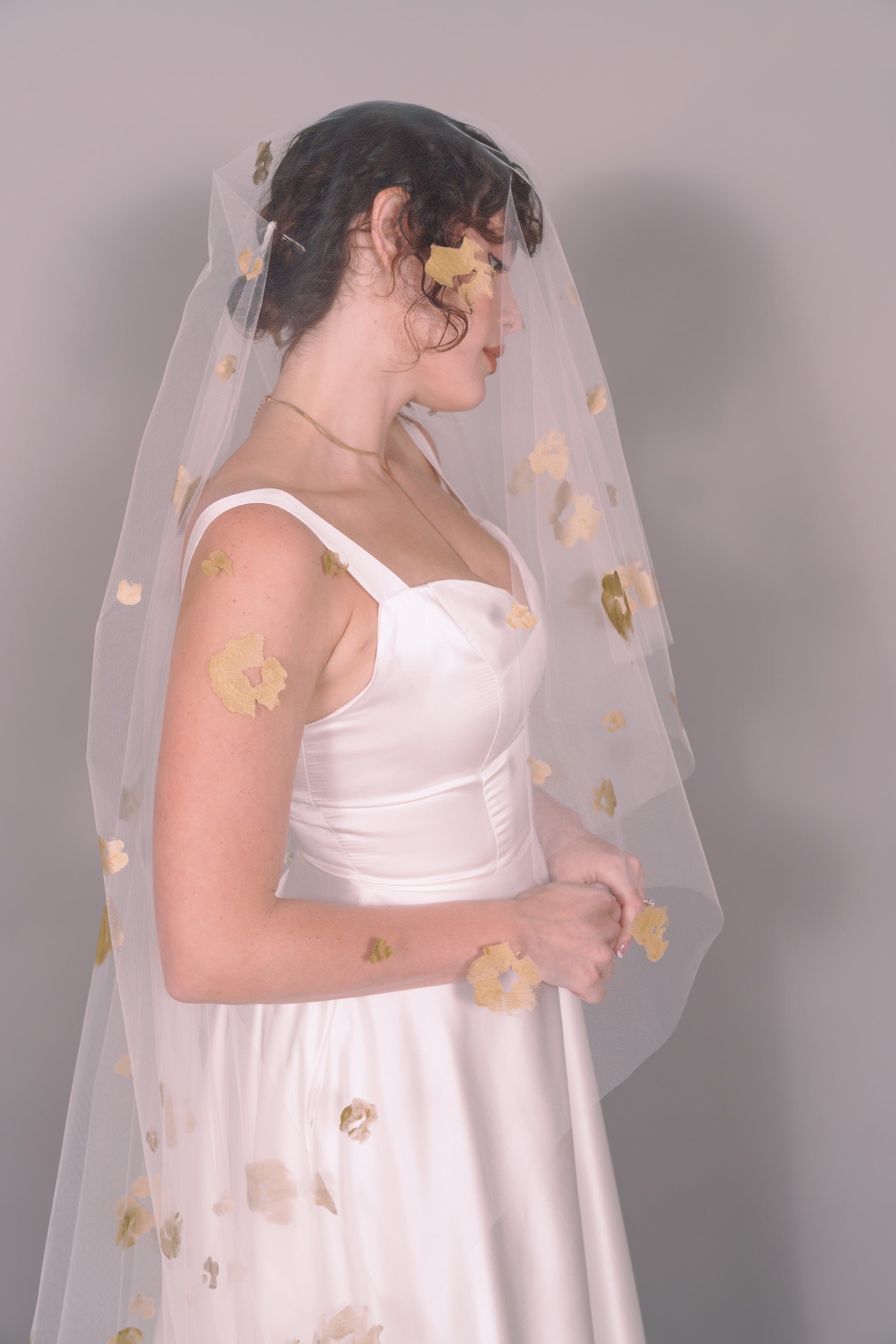 Woman wearing a wedding dress and ivory veil with painted gold floral petals on a plain background. Photography by Rachel Sakakeeny Model is Isabelle Paulino