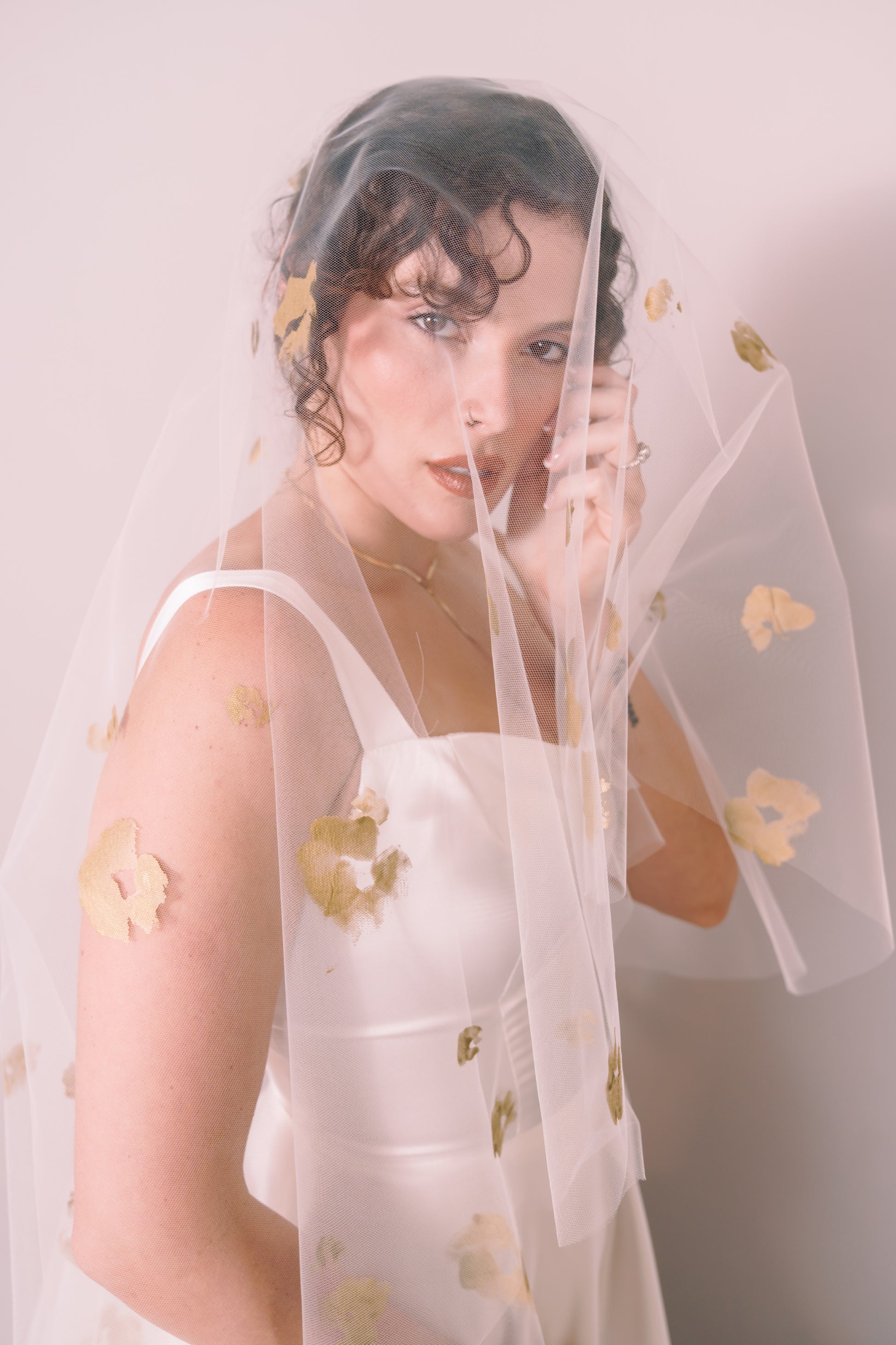 Woman wearing a wedding dress and ivory veil with painted gold floral petals on a plain background. Photography by Rachel Sakakeeny Model is Isabelle Paulino