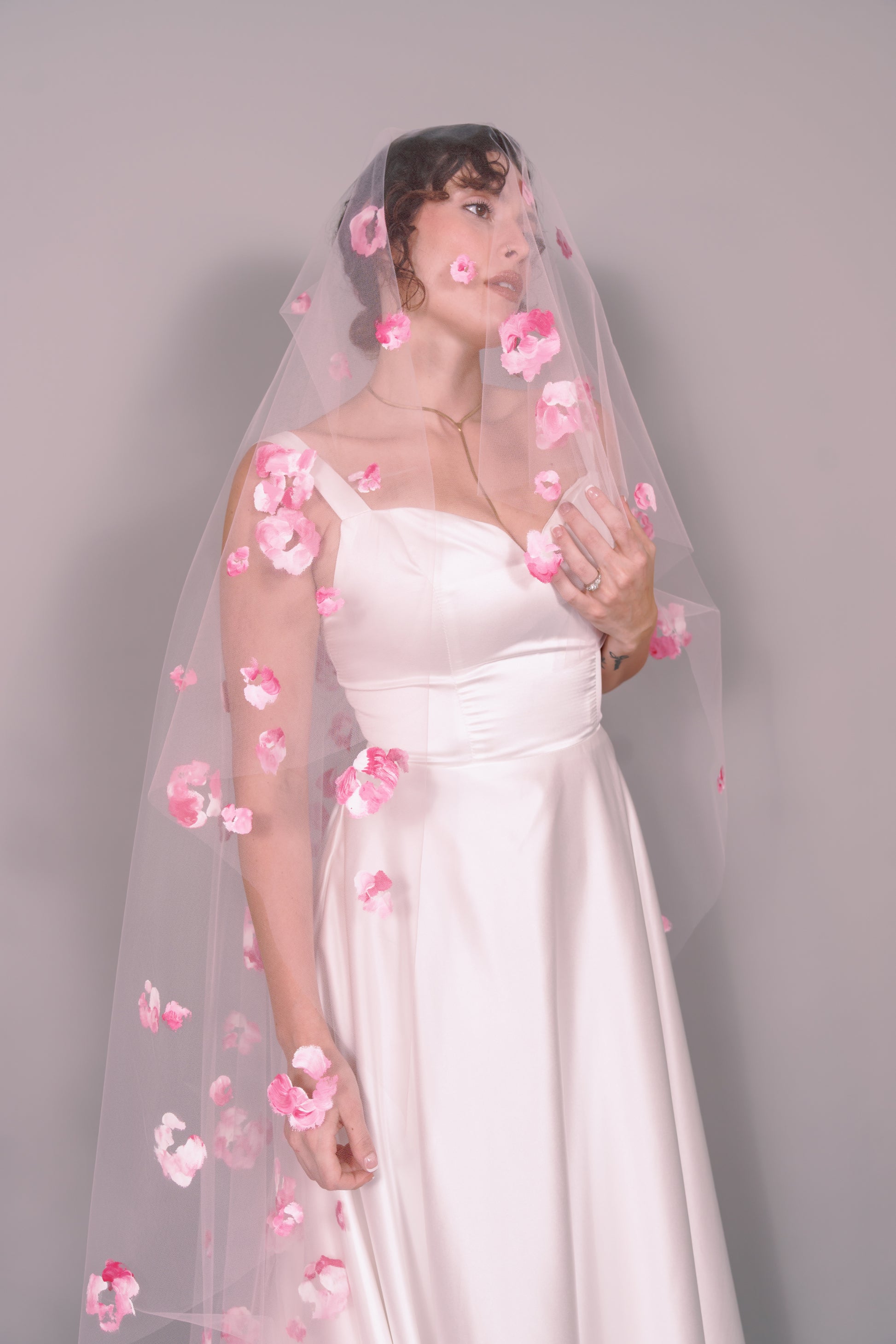 Woman wearing a wedding dress and pink veil with painted pink floral petals on a plain background. Photography by Rachel Sakakeeny Model is Isabelle Paulino