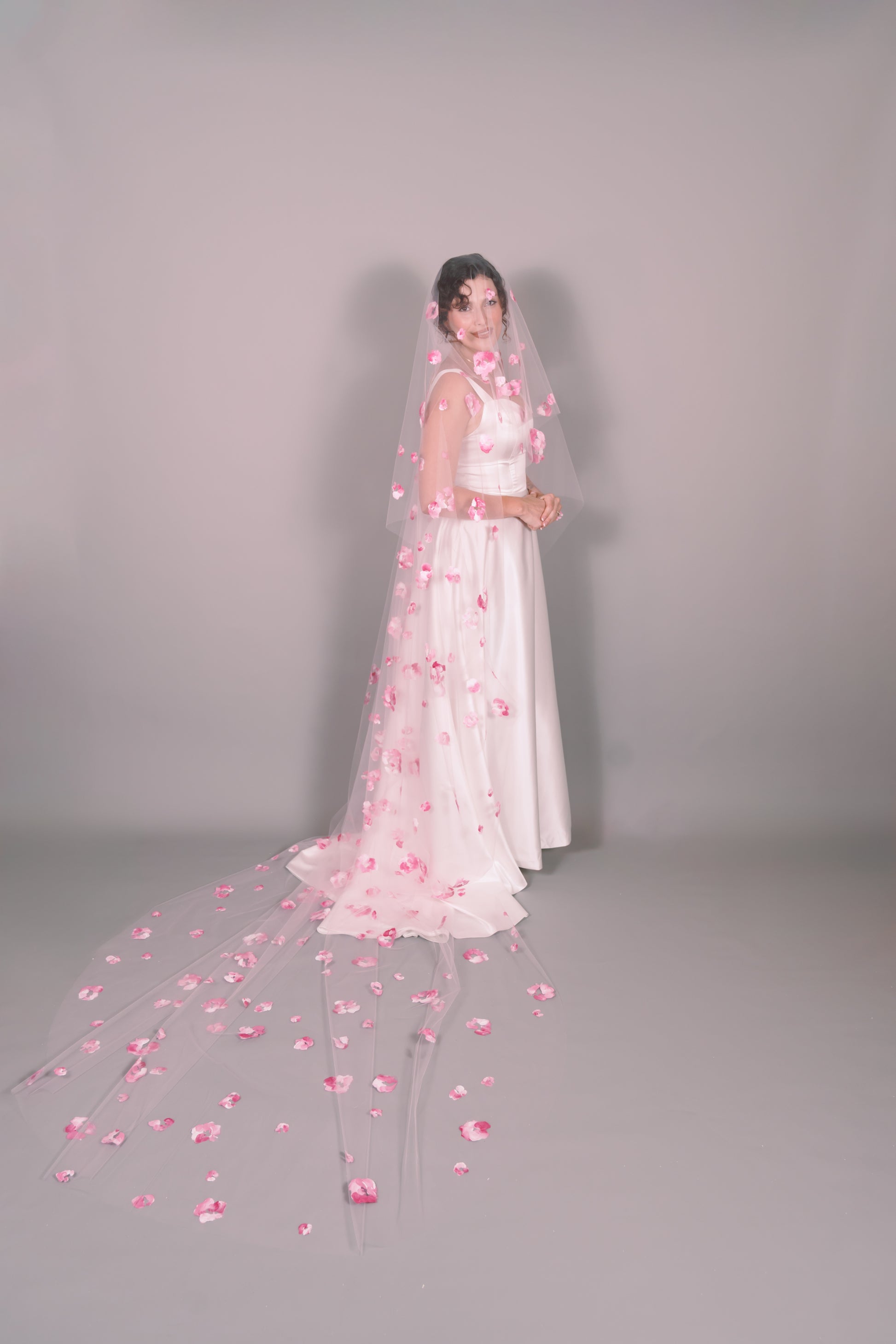 Woman wearing a wedding dress and pink veil with painted pink floral petals on a plain background. Photography by Rachel Sakakeeny Model is Isabelle Paulino