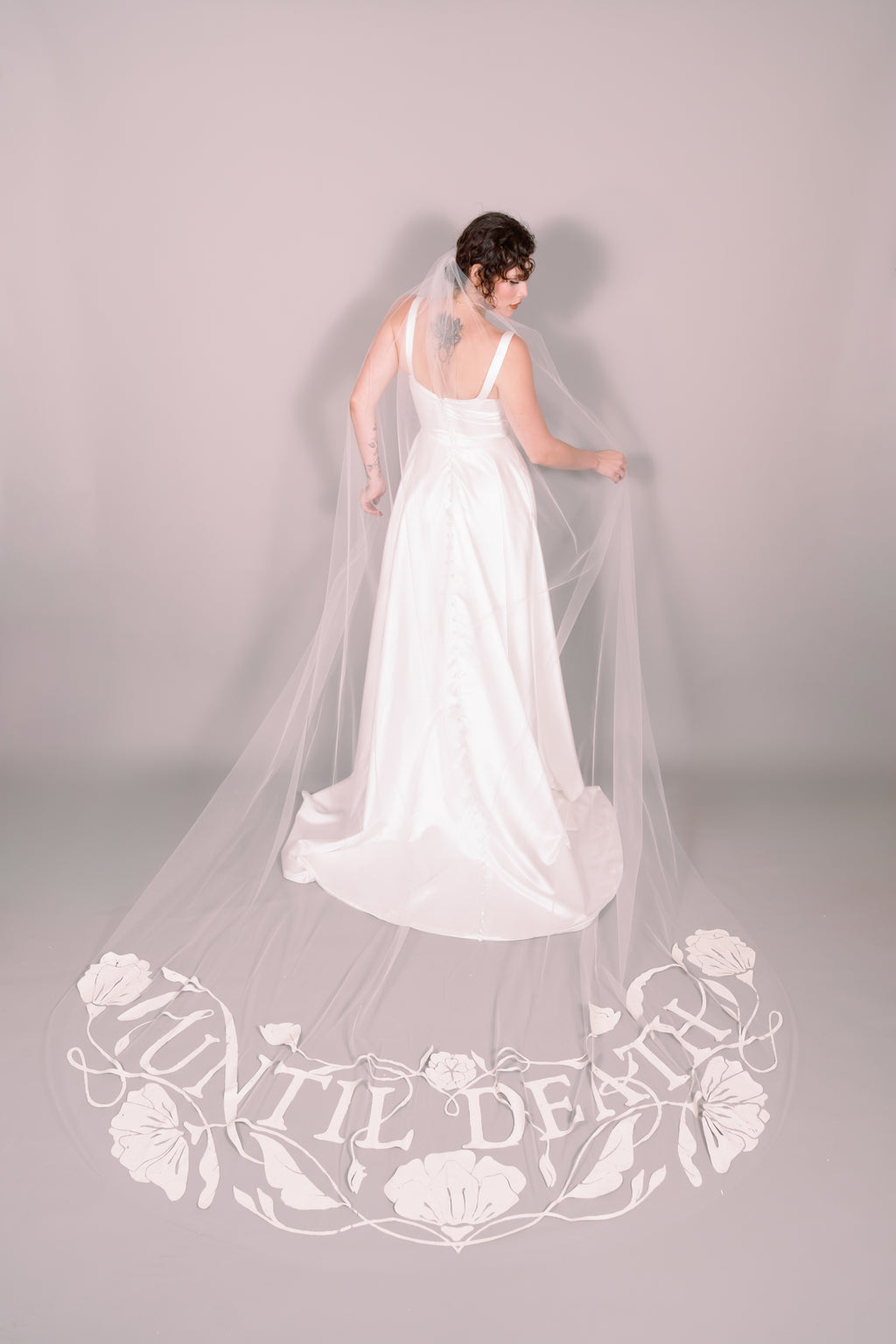Woman in a white wedding dress with a long painted veil that says Until Death against a plain background. Photography by Rachel Sakakeeny Model is Isabelle Paulino