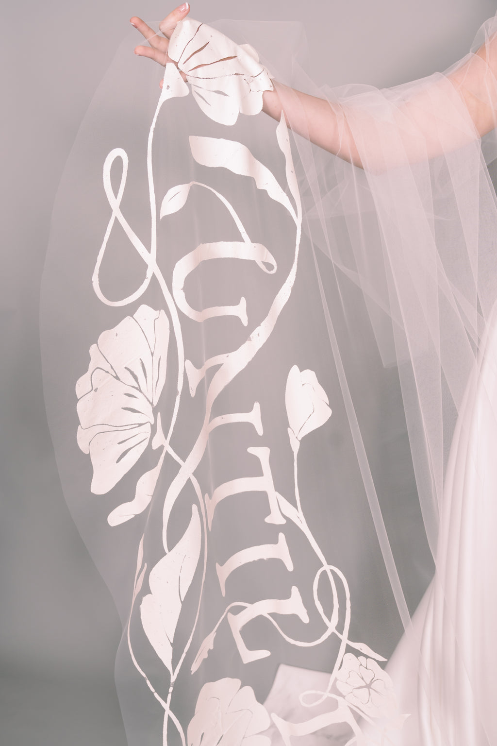 Veil with floral and leaf patterns, held by a hand against a neutral background. Painted veil says Until Death. Photography by Rachel Sakakeeny Model is Isabelle Paulino